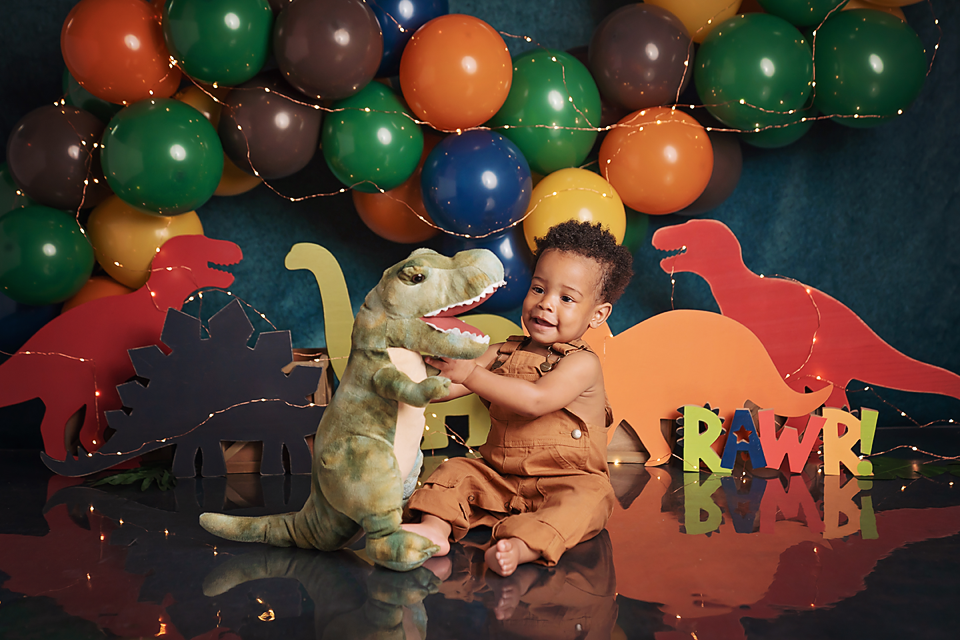 Baby boy in dinosaur themed first birthday photo with colorful balloons, dino cutouts, and plush T-Rex toy.