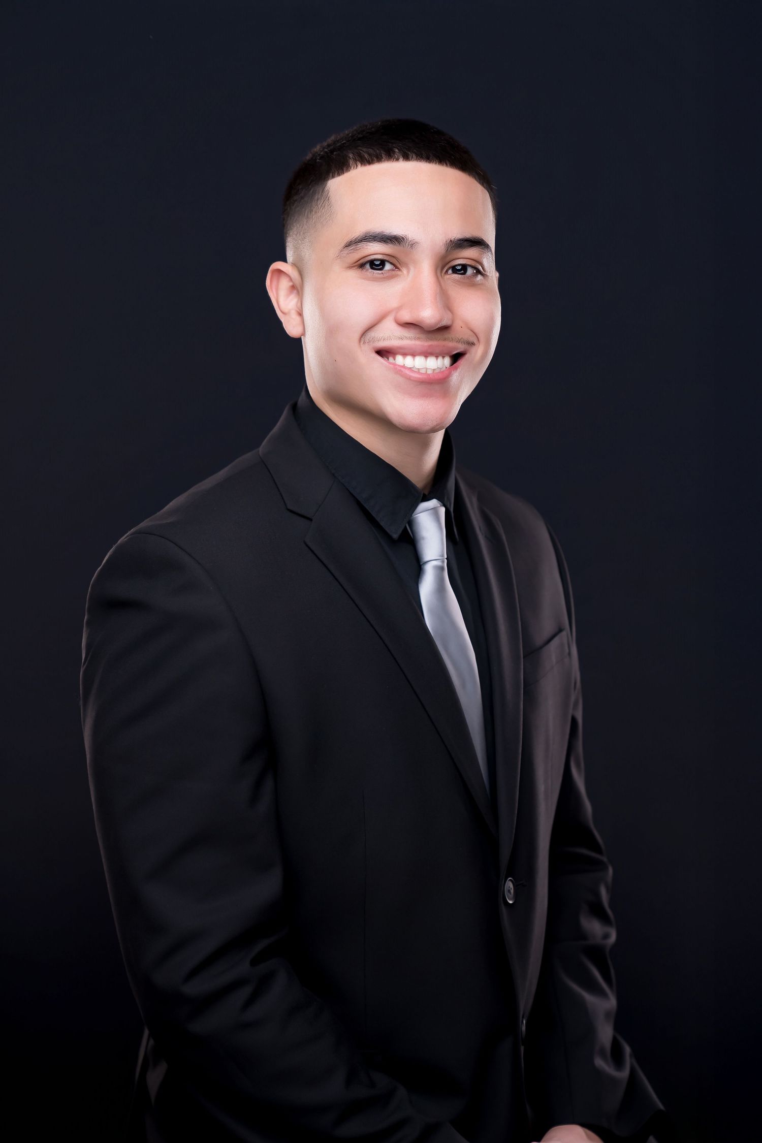 Young man in black suit and silver tie smiling confidently against dark background.