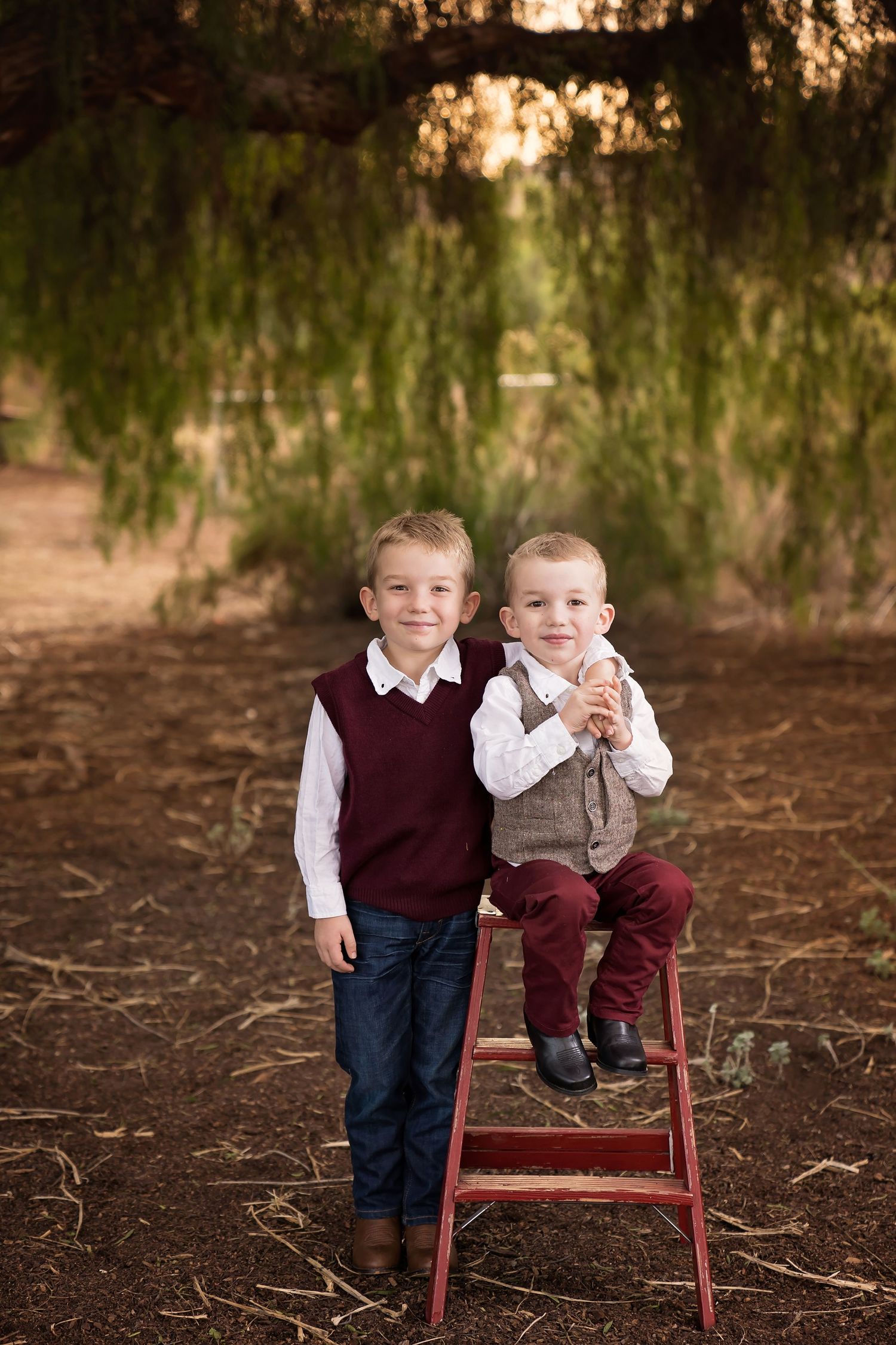 Two children in coordinating outfits posing with a red ladder beneath dreamy willow trees — San Diego outdoor family session