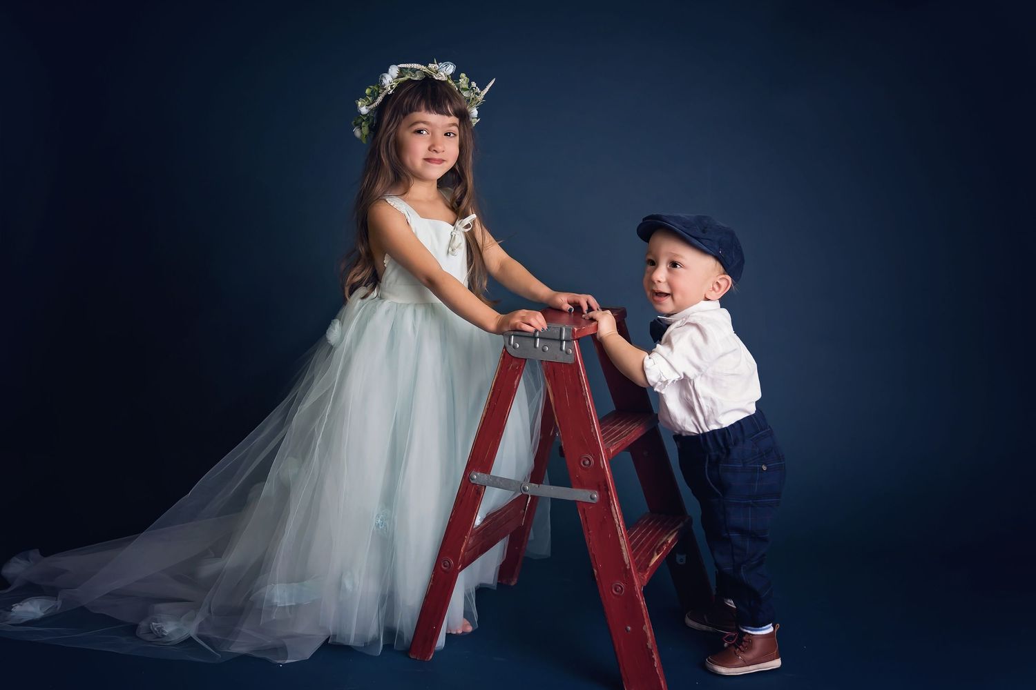 Children in formal wear pose playfully with red ladder against dark studio backdrop.