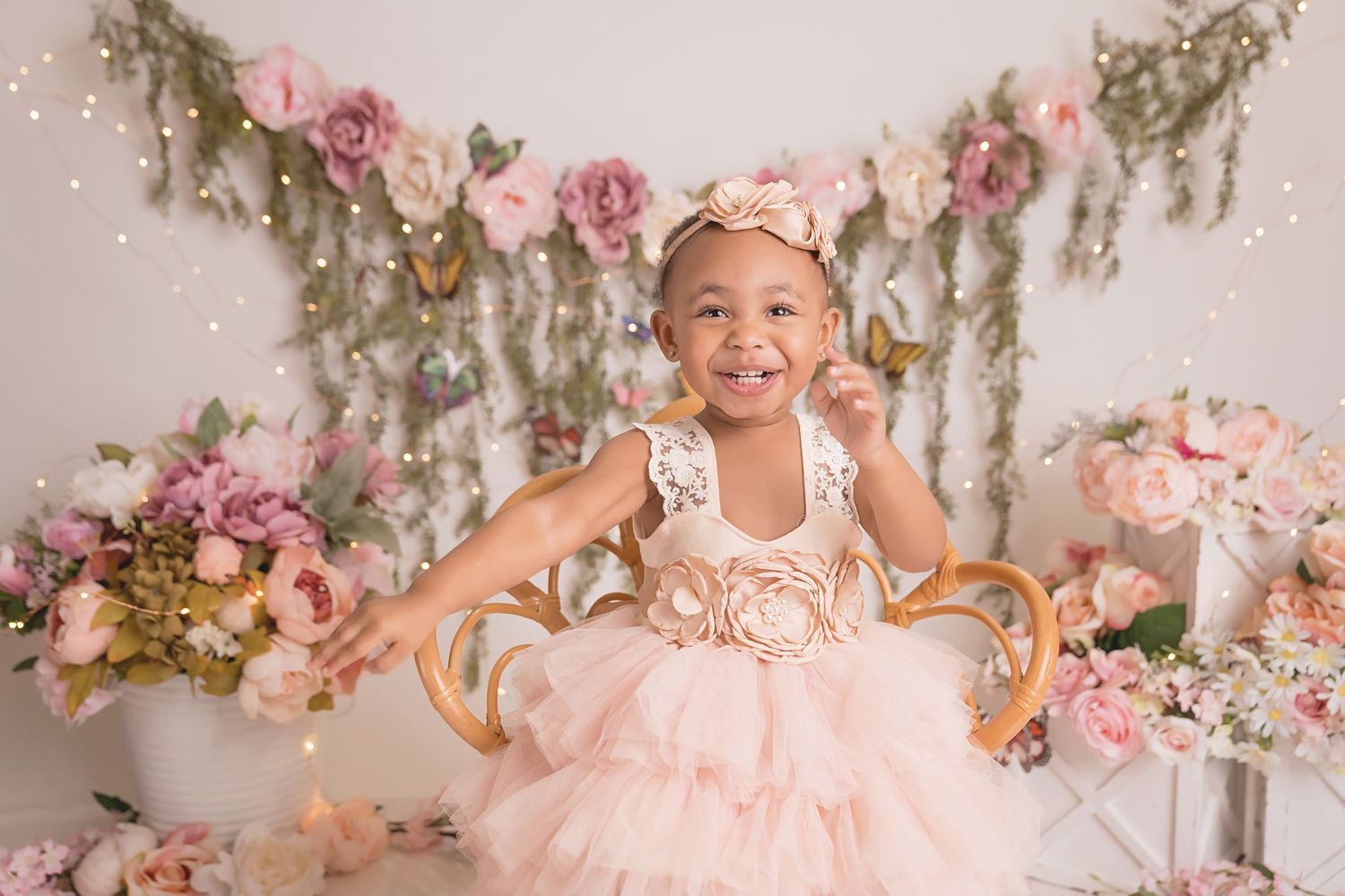 Child in pink tulle dress posing joyfully on chair with floral and butterfly backdrop.