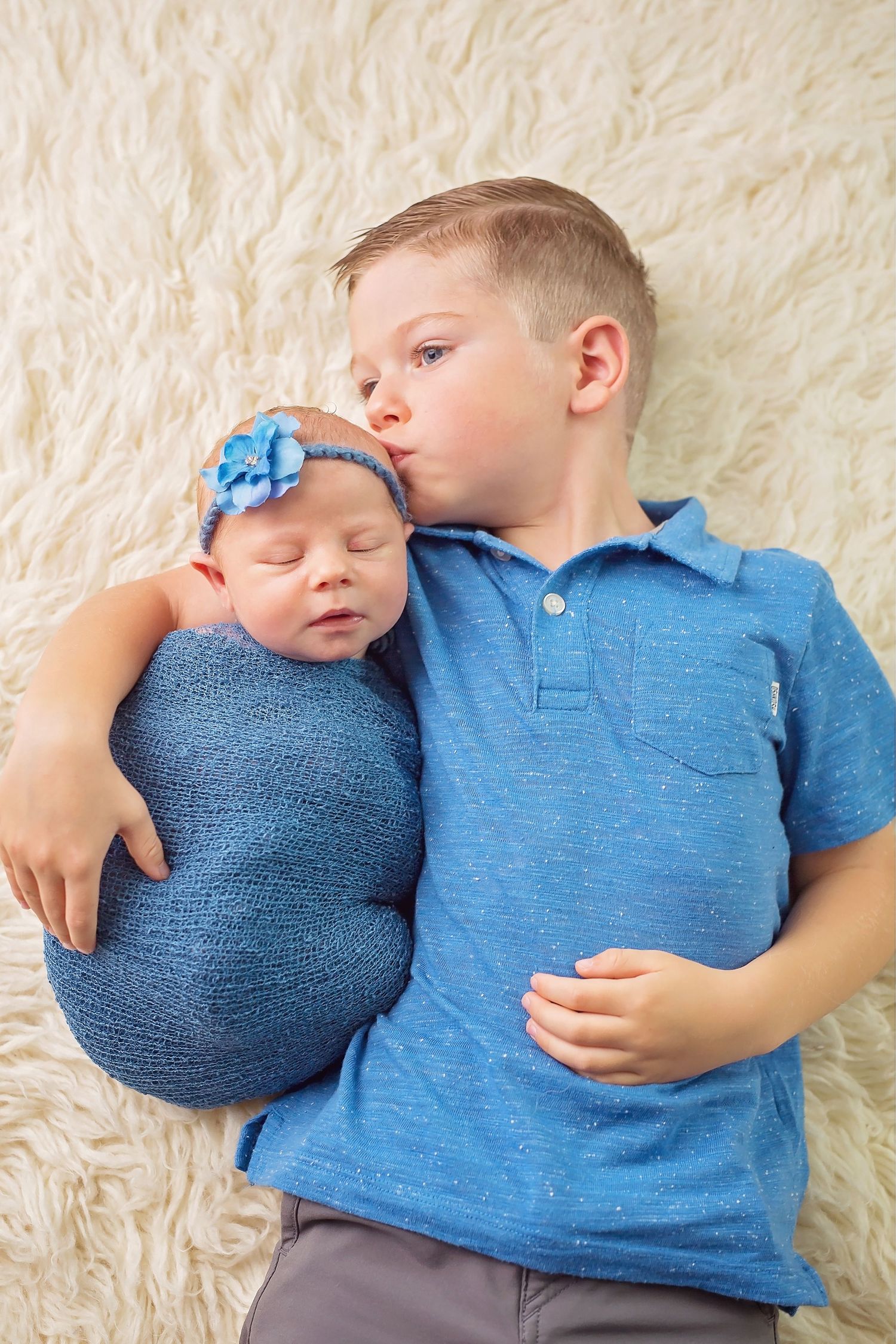 Siblings in matching blue outfits lying together on white fluffy rug. San Diego Newborn Photographer