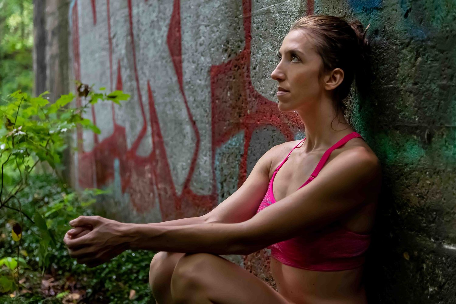 A fit woman in a pink sports bra sits against a graffiti-covered concrete wall surrounded by lush green foliage.