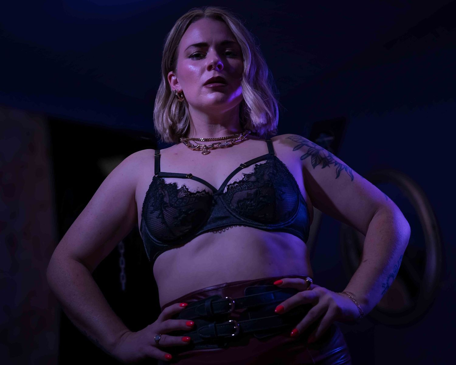 Young woman with tattoos posing confidently in black lace lingerie under blue stage lighting.