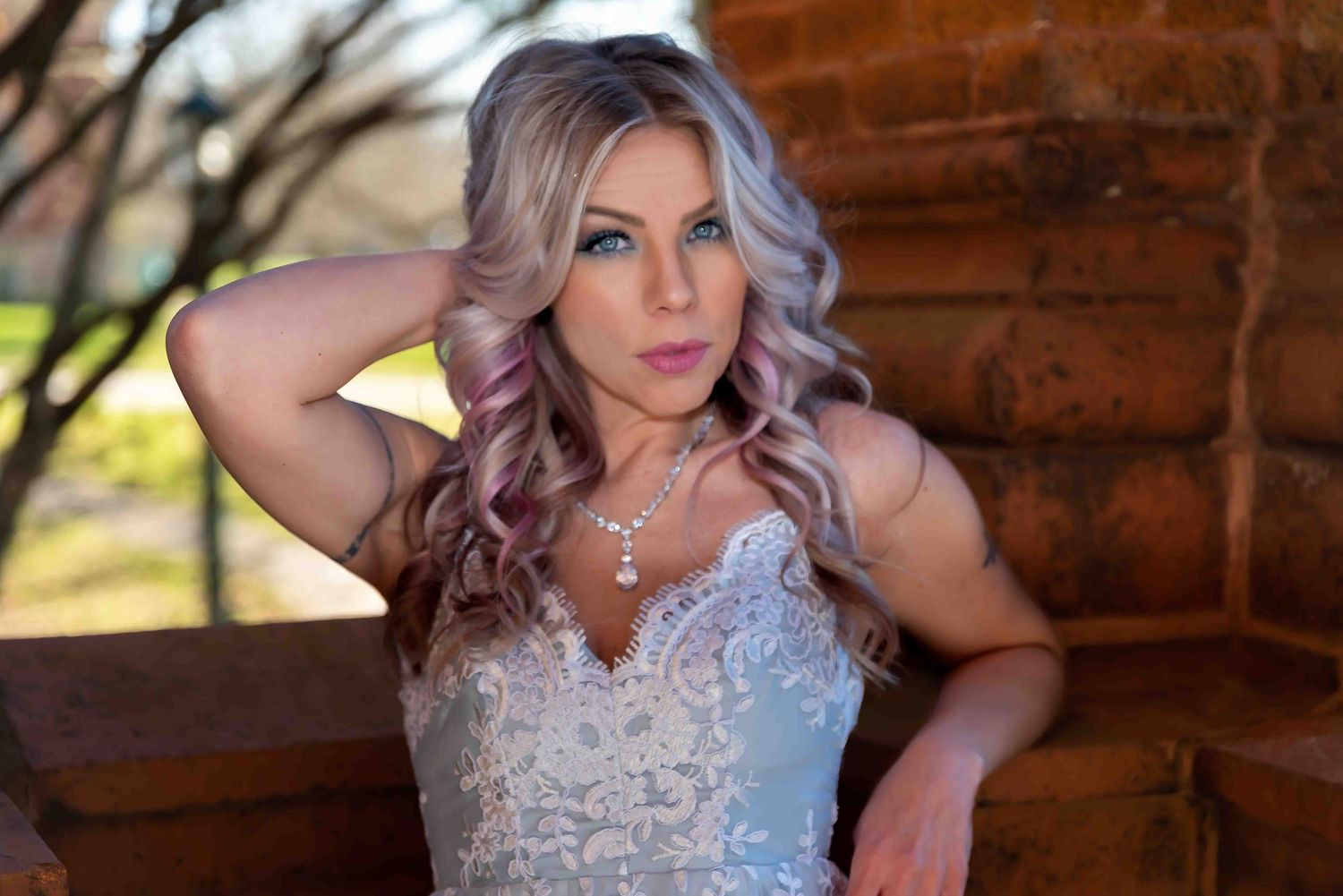 Woman in white lace gown with pink-highlighted hair poses gracefully near rustic log wall in elegant bridal portrait.