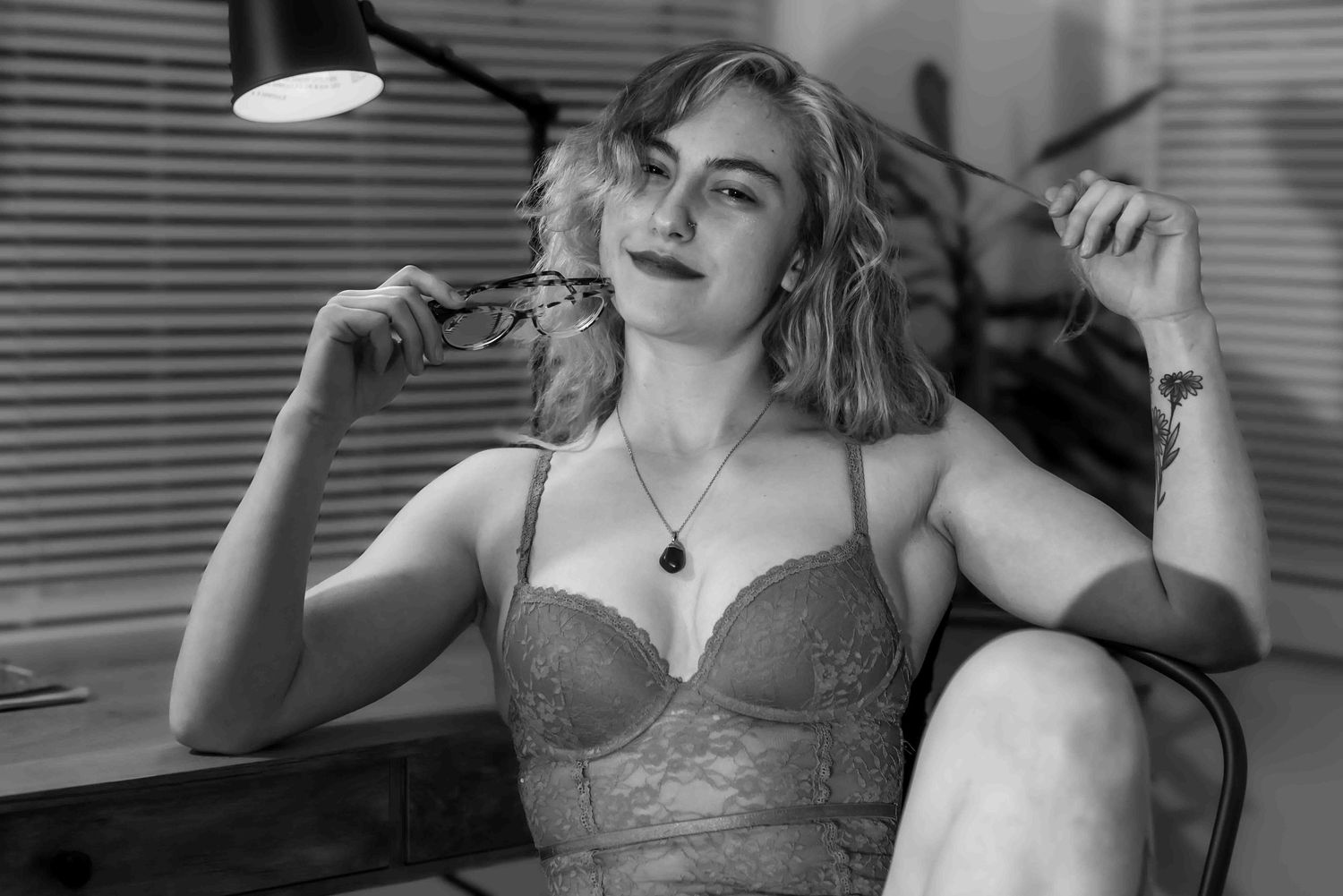 Black and white portrait of a woman in lace bra holding glasses while seated at a desk.