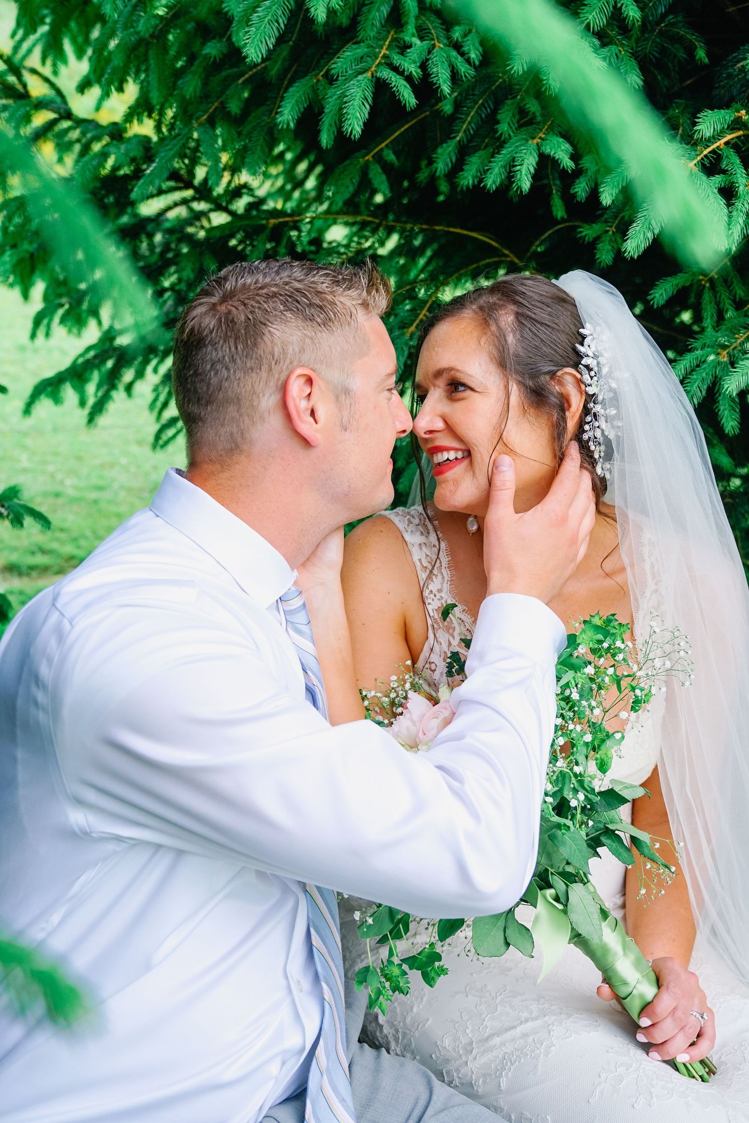 A romantic wedding moment captured under lush green foliage with a bride in white and groom in light attire.