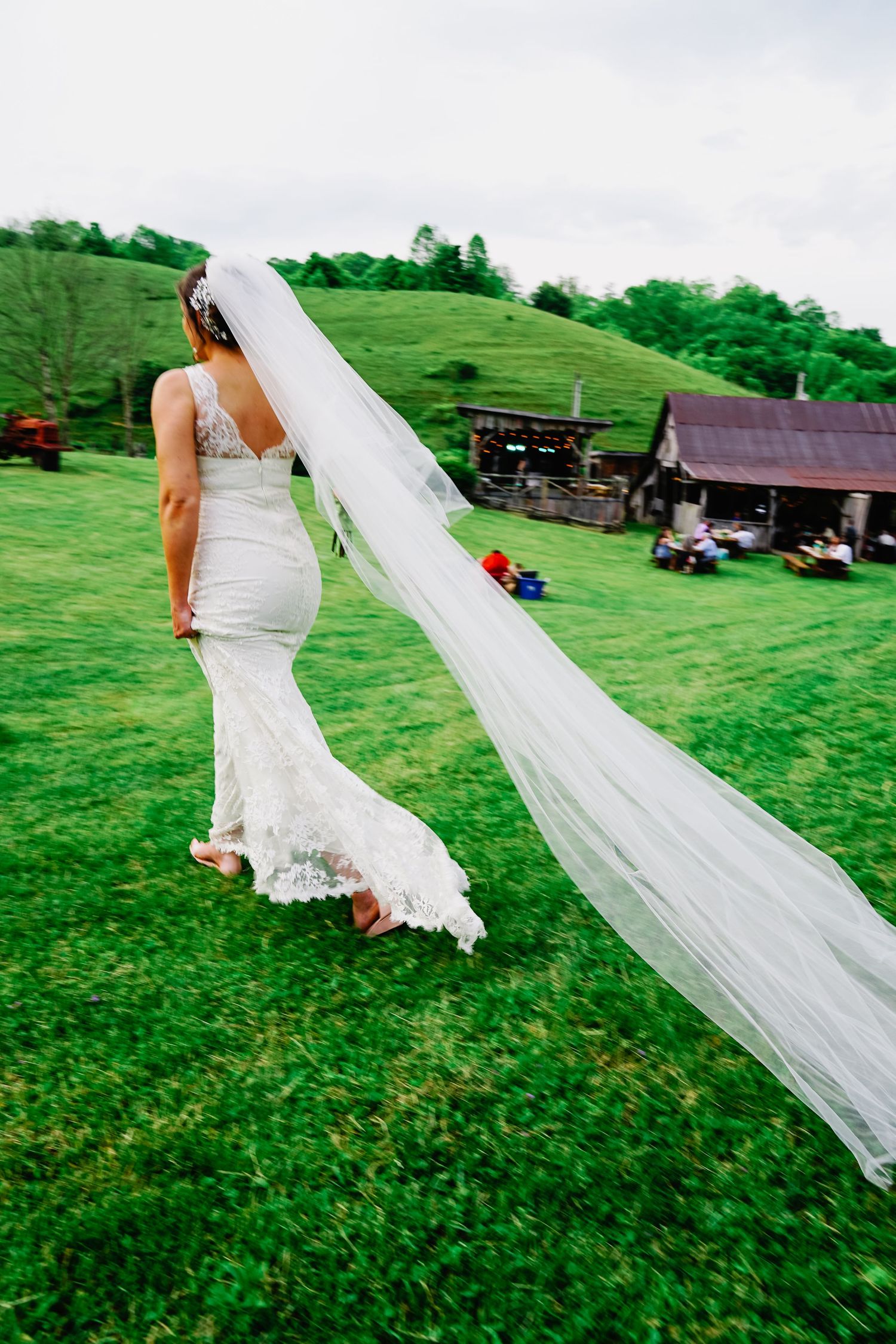 A flowing white wedding veil trails behind a bride walking across a lush green hillside on a sunny day.