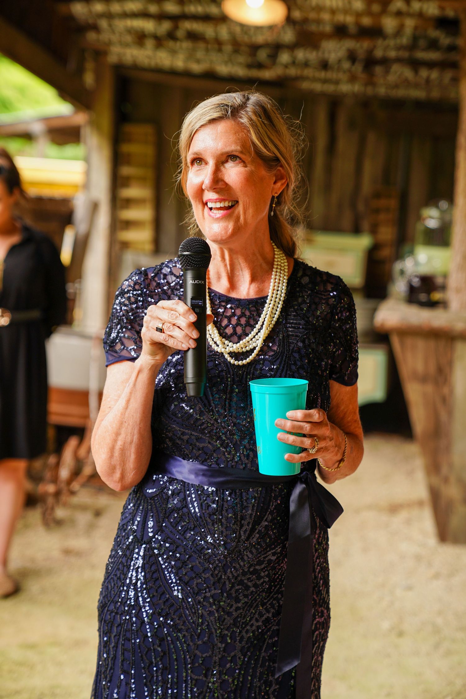 Someone in a sparkly navy blue dress holds a turquoise drink cup while laughing outdoors at a social gathering.