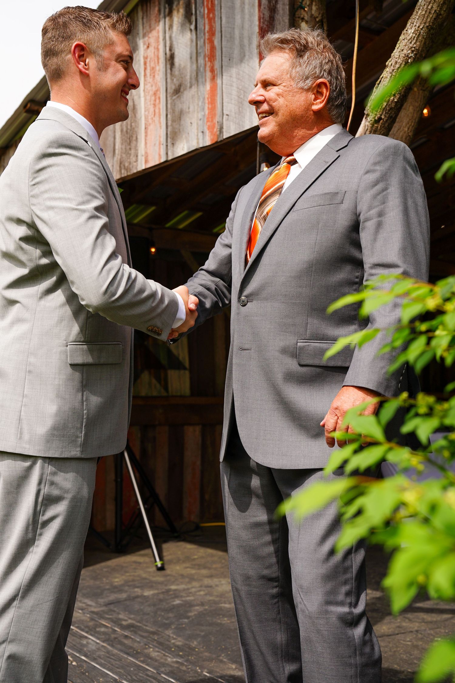 Two people in formal suits shake hands and share a friendly moment in front of a rustic wooden backdrop.