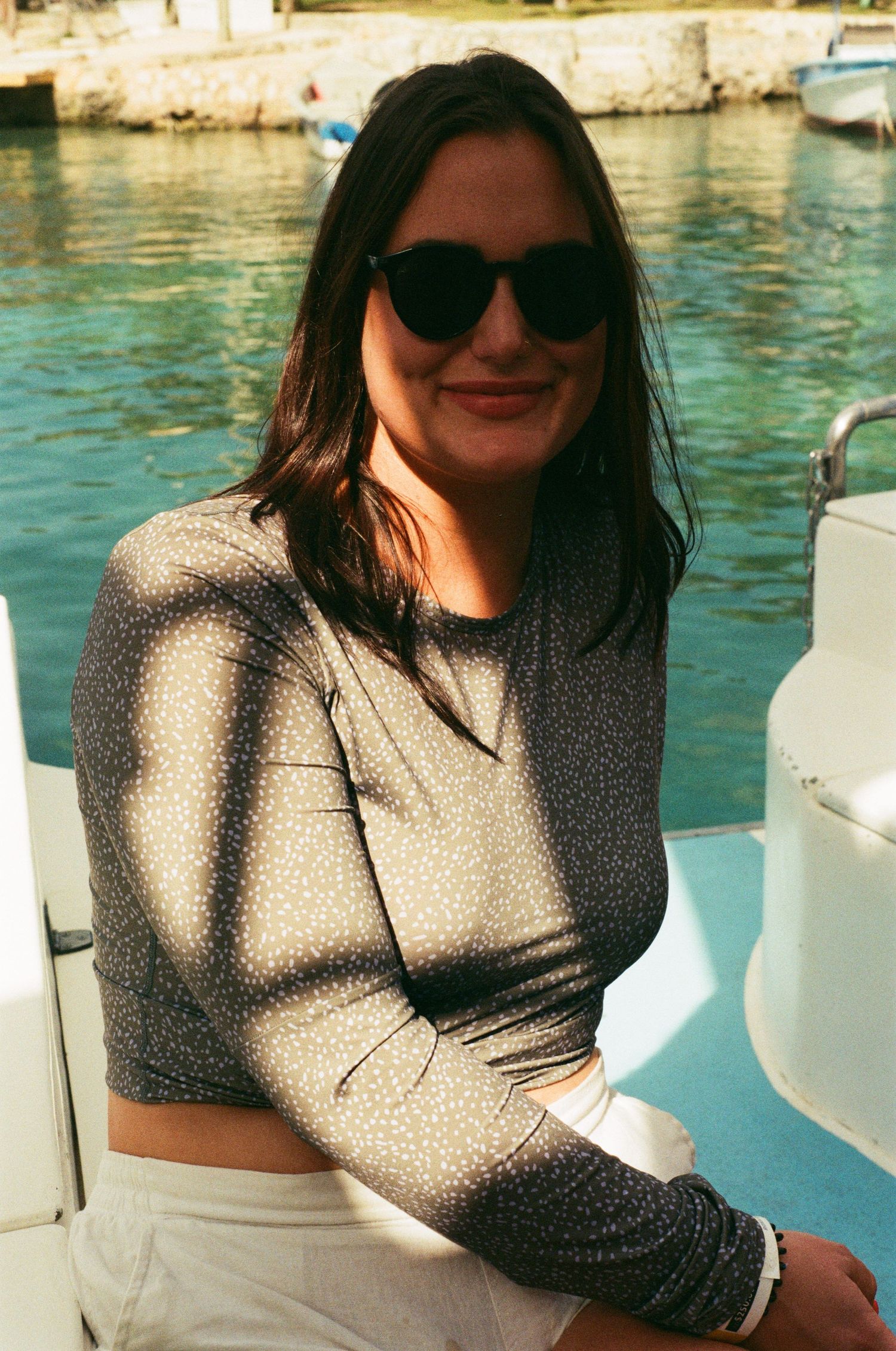 A person wearing sunglasses and a sun shirt sits by a marina with turquoise water and boats in the background. 35mm film