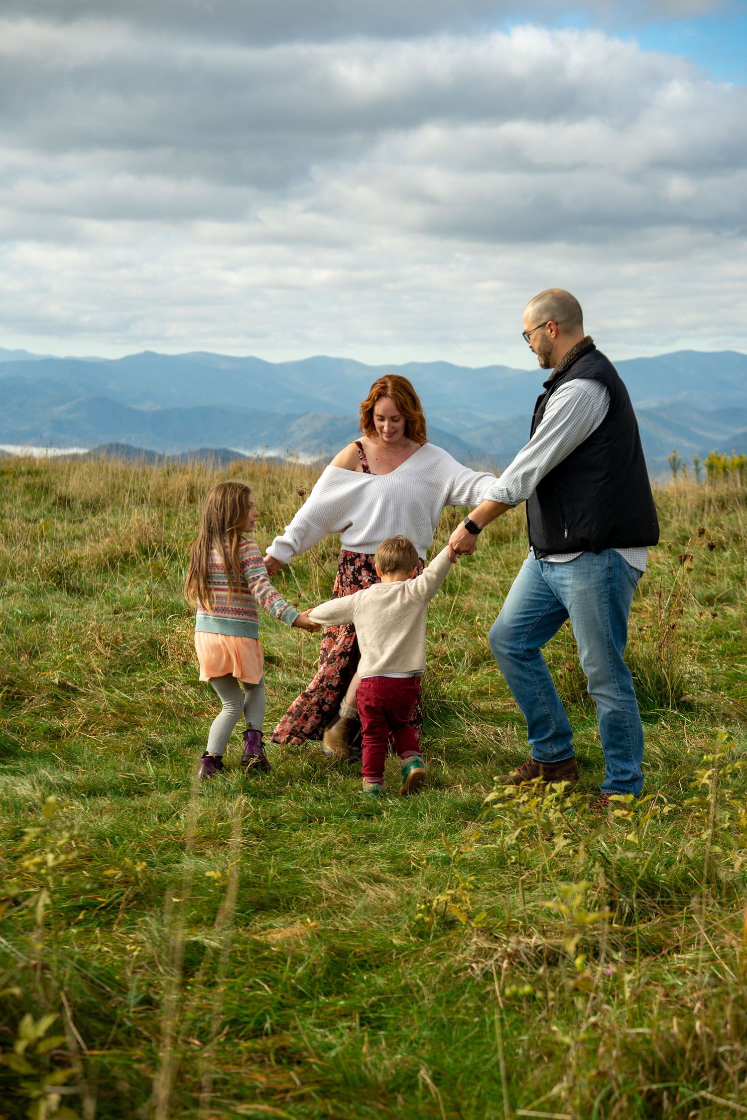 A family explores a grassy hilltop meadow with mountains visible in the cloudy background.