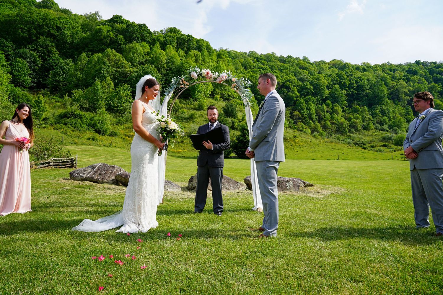 Wedding ceremony taking place outdoors on a lush green hillside with guests gathered around on a sunny day.