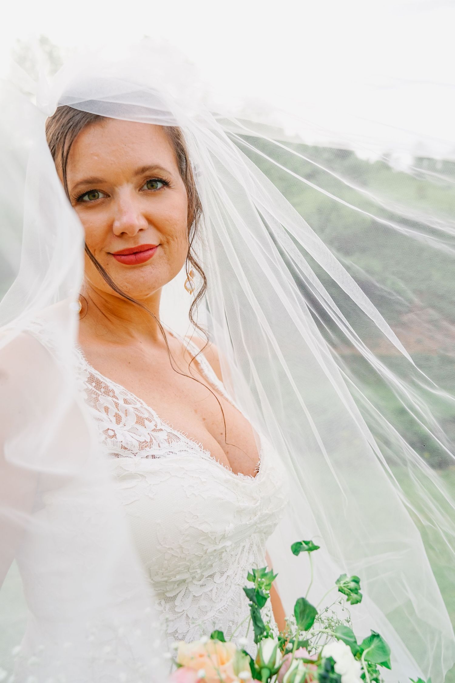A bride wearing a white lace wedding dress and flowing veil holds a bouquet with peach roses and greenery.