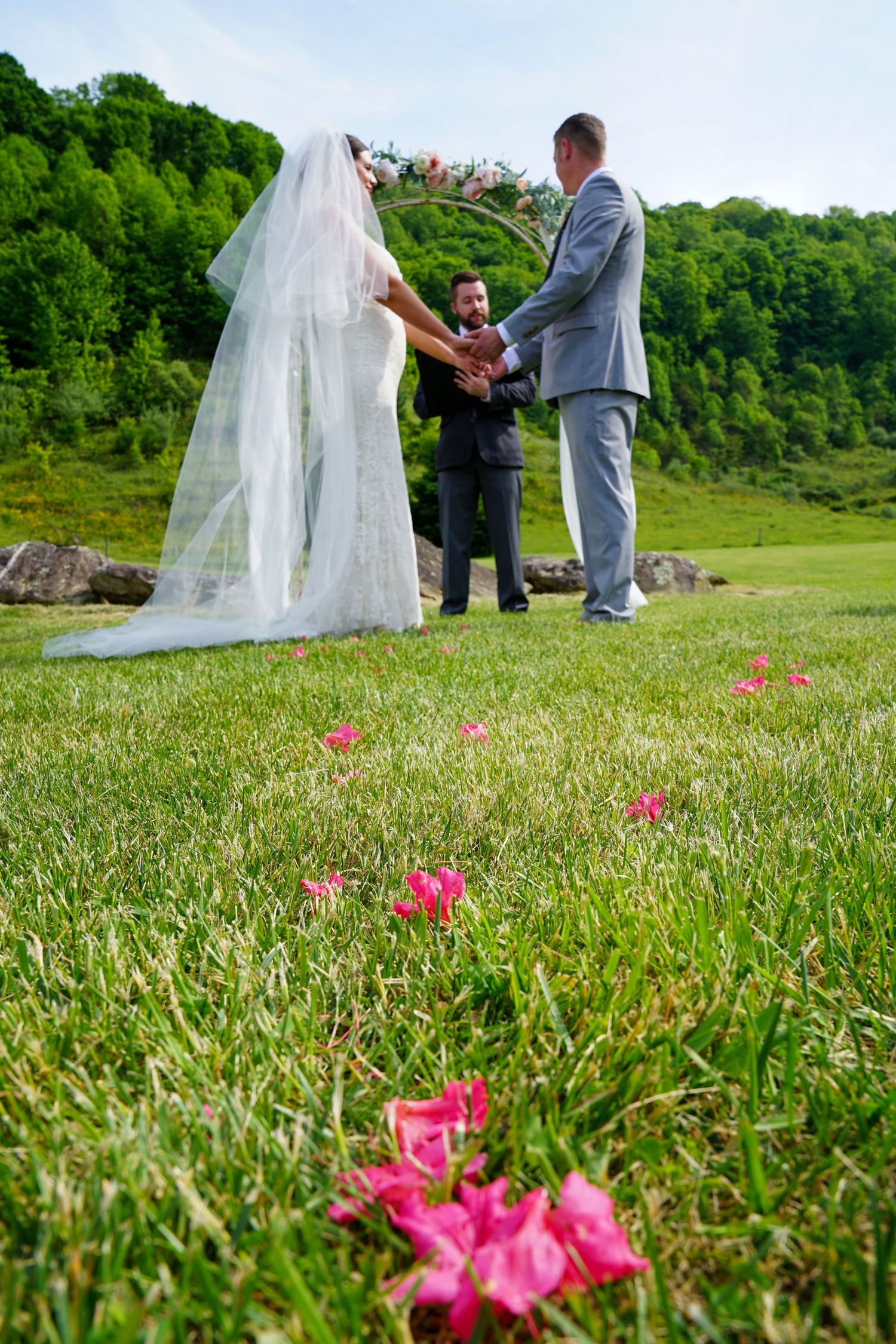 A wedding ceremony takes place on a grassy hillside with bright pink flowers in the foreground.