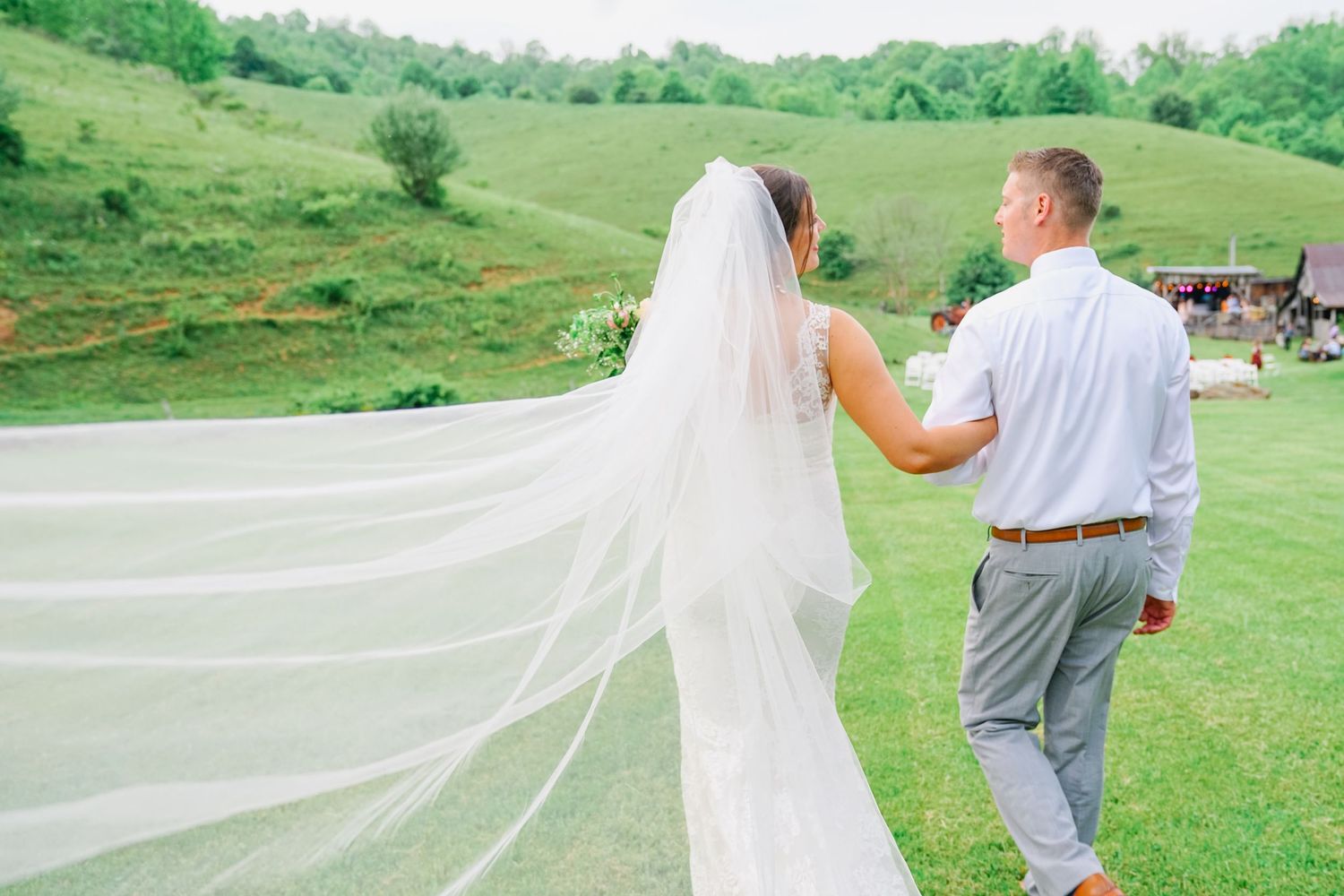 A long flowing bridal veil trails behind a couple walking on a green hillside during an outdoor wedding ceremony.