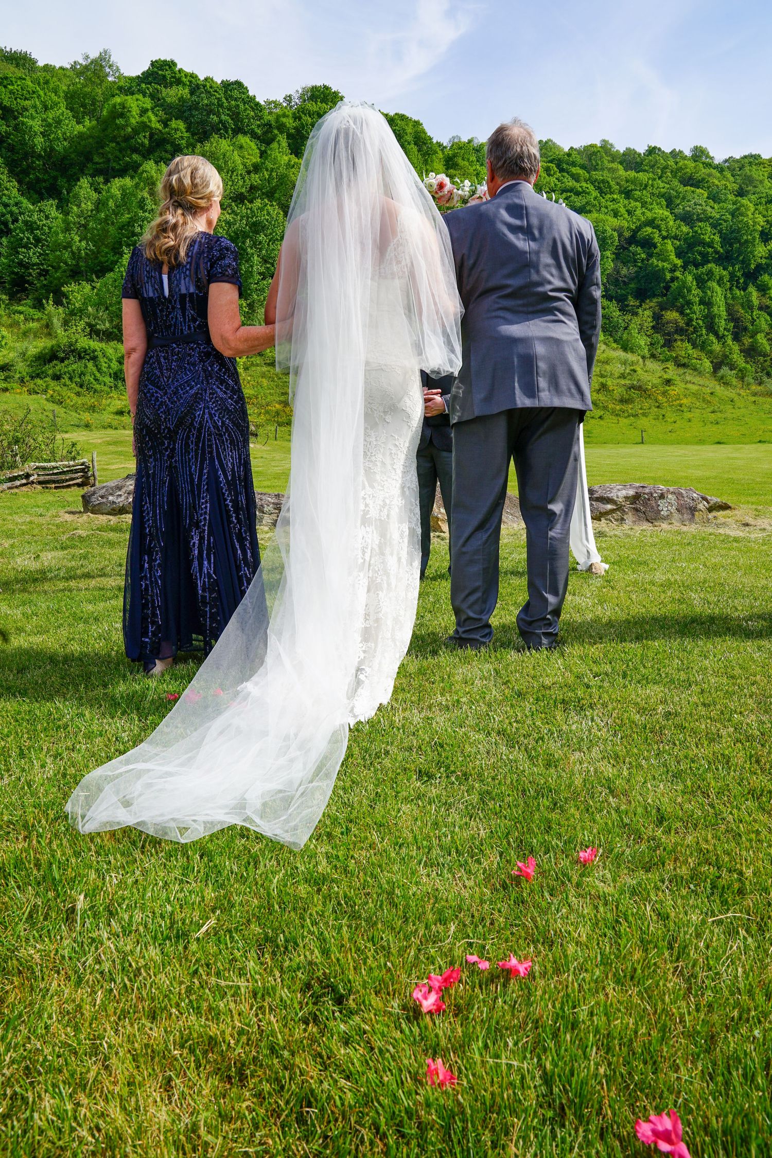 Wedding ceremony participants stand in a green field with pink flowers as a flowing white veil trails behind them.