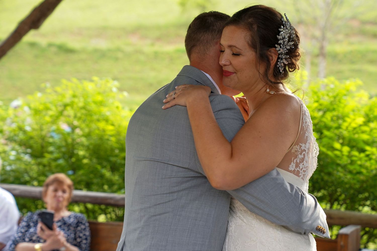 A tender moment between newlyweds sharing their first dance at an outdoor wedding reception with greenery in background.