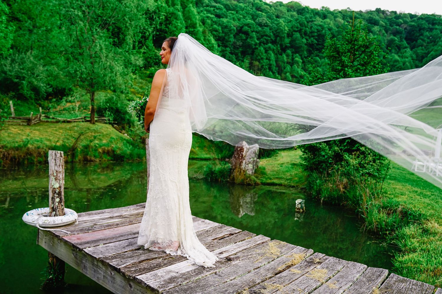 Long flowing veil catches the wind as someone poses on a rustic wooden bridge surrounded by lush green forest.