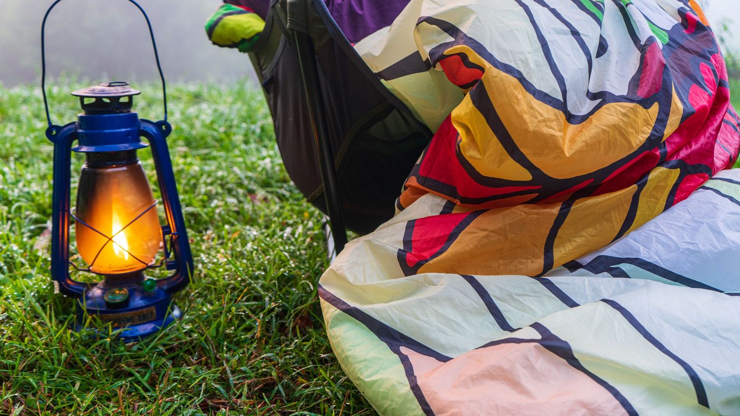 A blue vintage lantern glows warmly next to colorful bedding on grass.