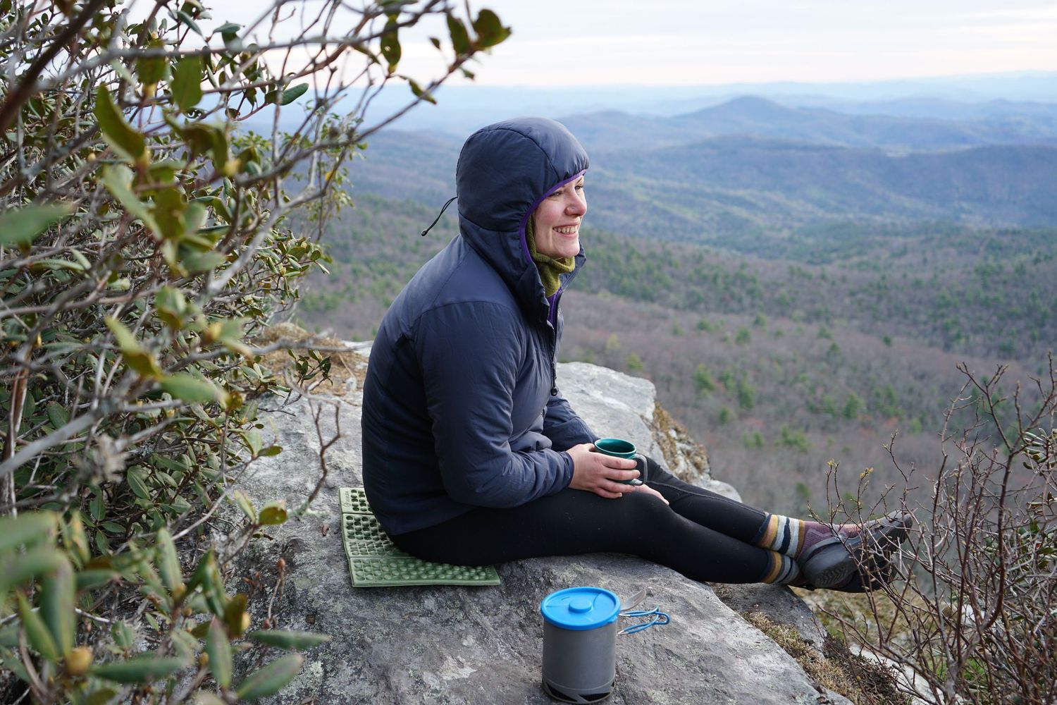 A person in dark clothing sits on a rocky cliff edge enjoying panoramic mountain views.