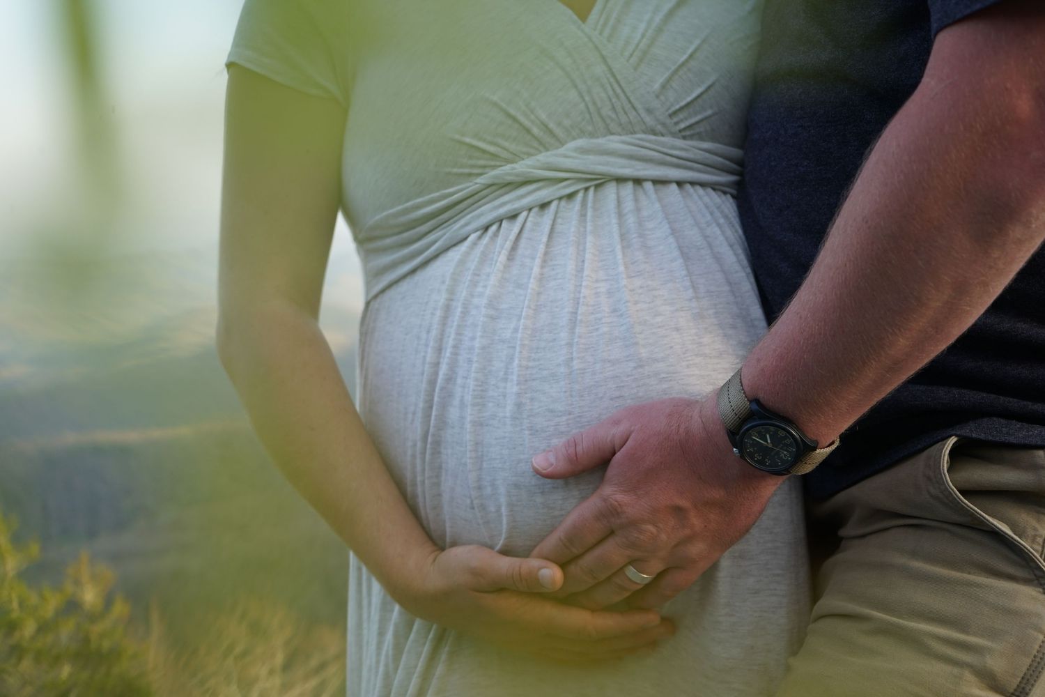Close-up of expectant parents holding hands over pregnant belly against mountain backdrop.