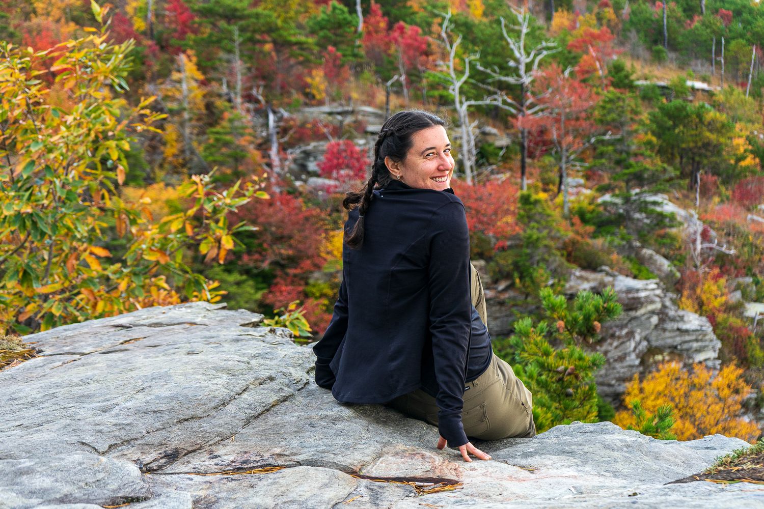 A hiker sits on a rocky cliff edge overlooking a scenic autumn mountain landscape with colorful foliage.