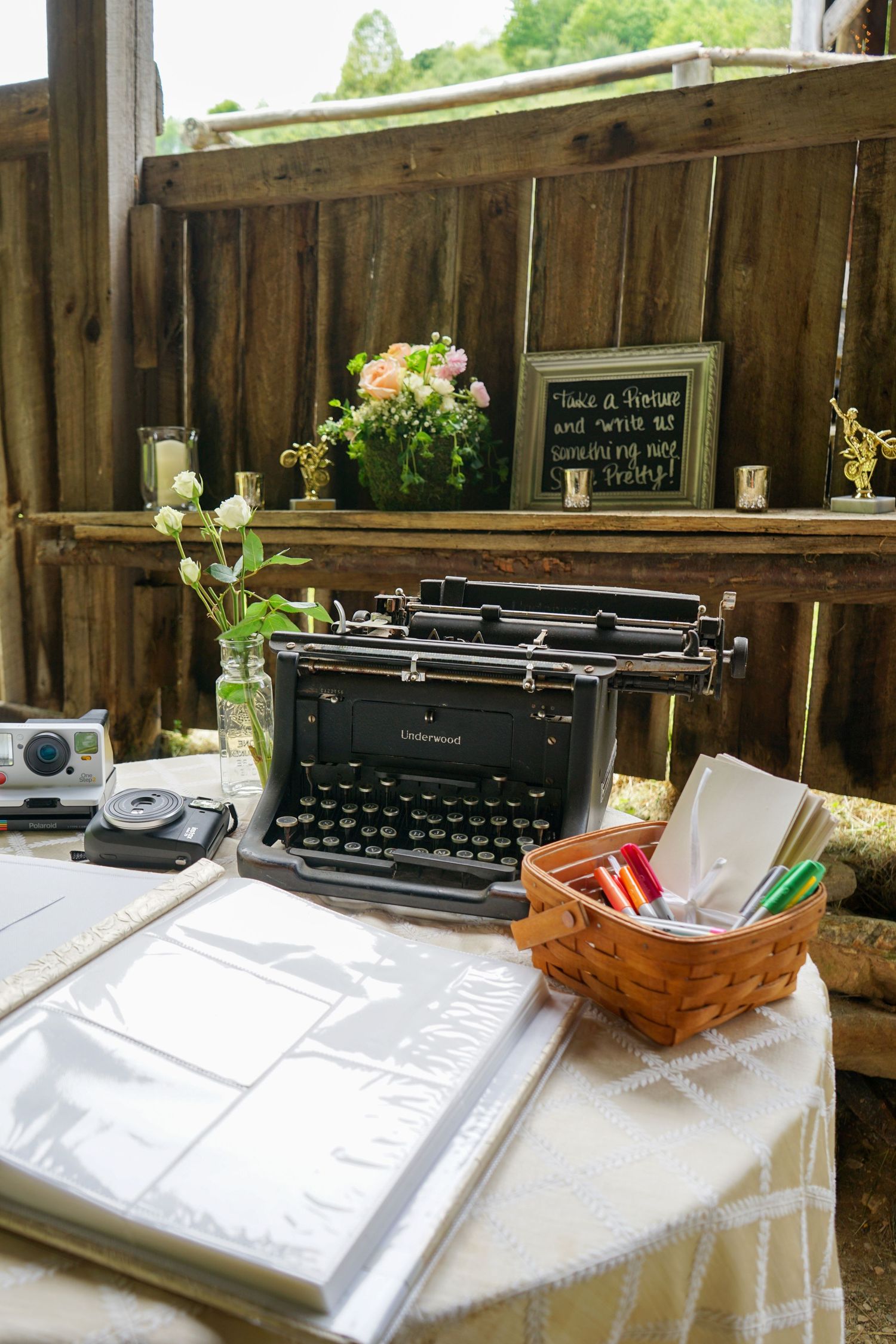 Vintage typewriter display on white tablecloth with wicker basket and floral decorations at rustic wooden venue.