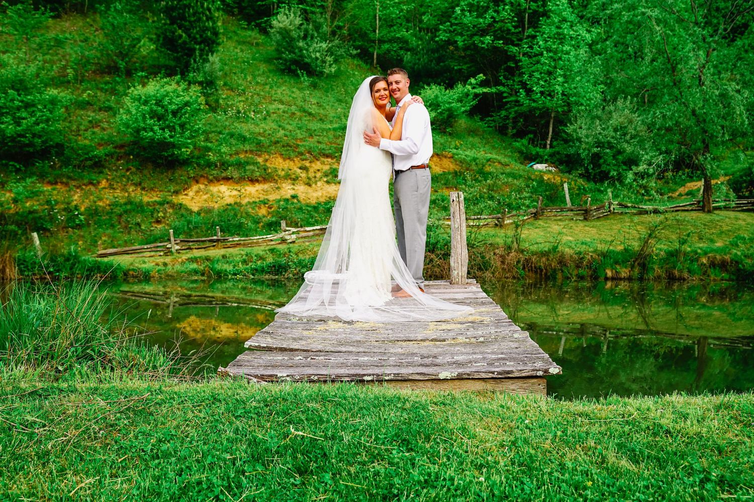 A couple in wedding attire embrace on a rustic wooden bridge over a pond surrounded by lush green landscape.