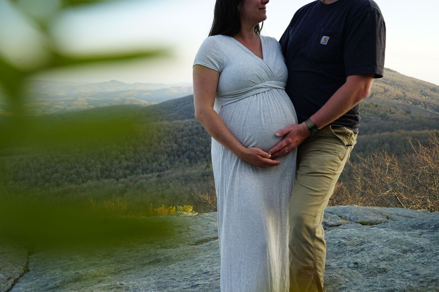 Expectant couple standing on rocky mountain overlook with scenic valley background.
