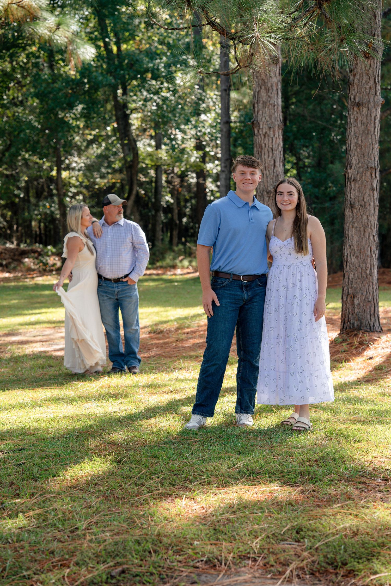 A couple stands together in a wooded area with sunlight filtering through pine trees on a sunny afternoon.