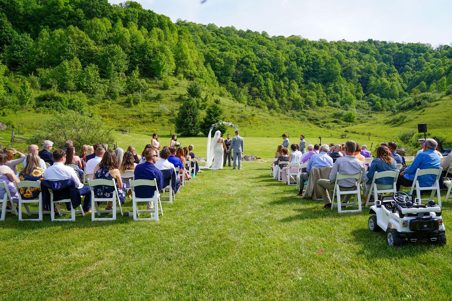 Guests seated in white chairs on a lush green hillside for an outdoor wedding ceremony.