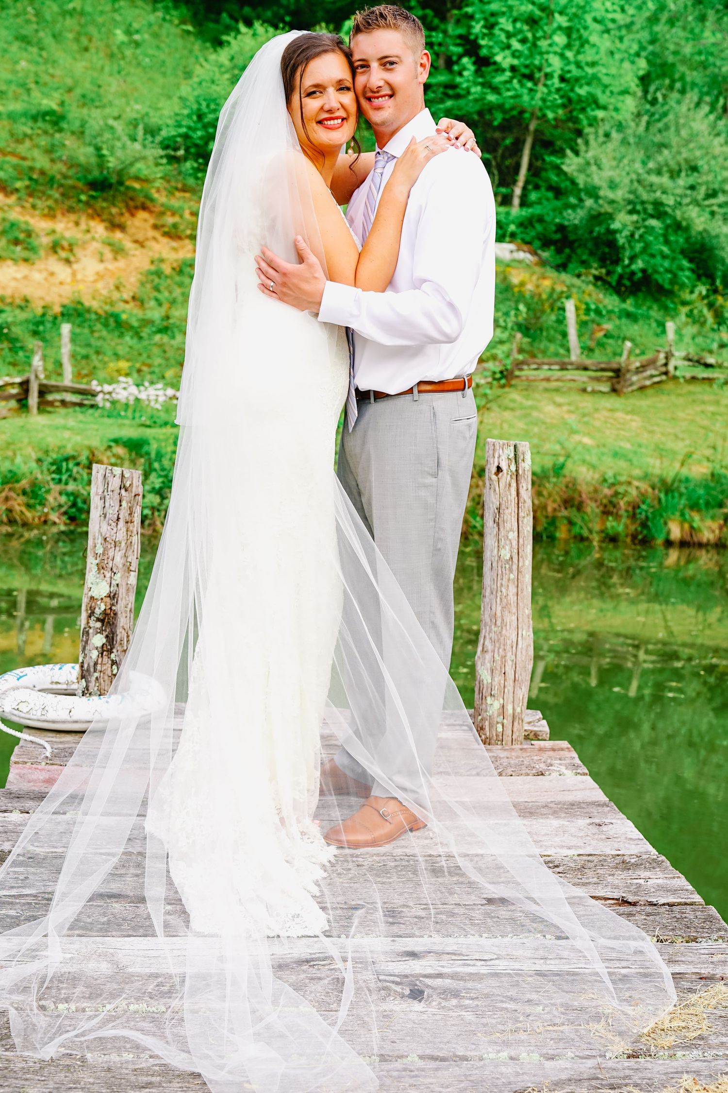 A couple shares romantic moments on a rustic wooden dock surrounded by lush green scenery and a tranquil pond.