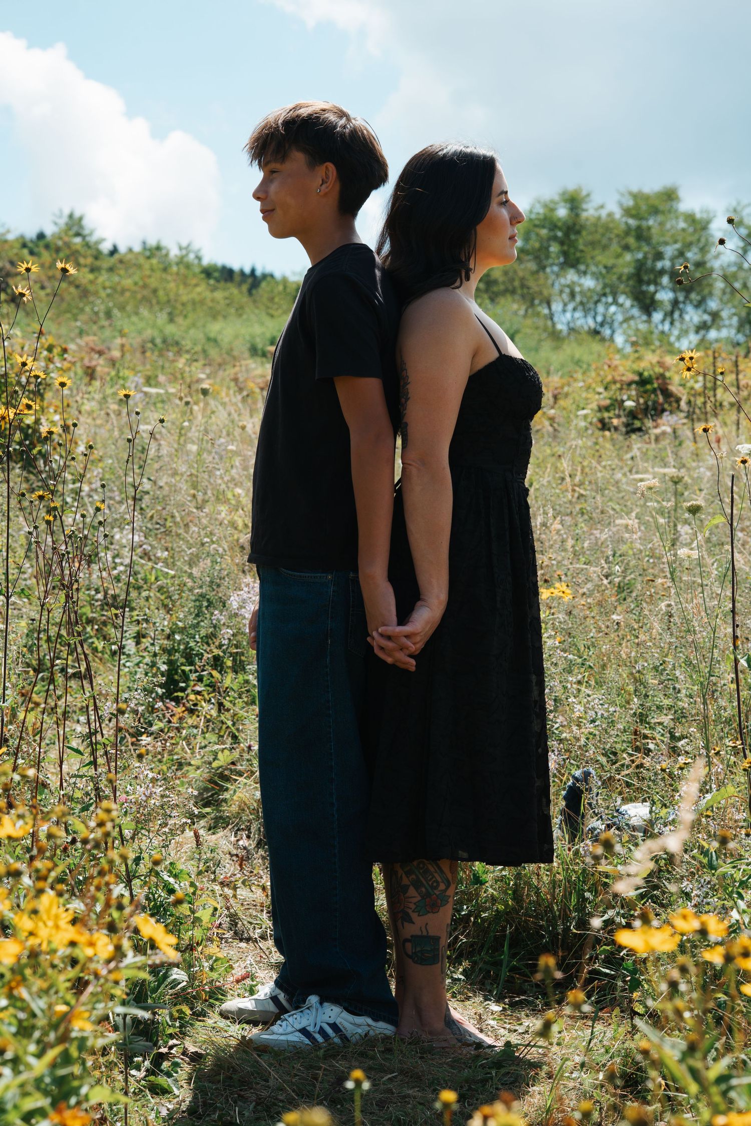 Two people stand close together in a wildflower meadow with yellow blooms and greenery on a sunny day.