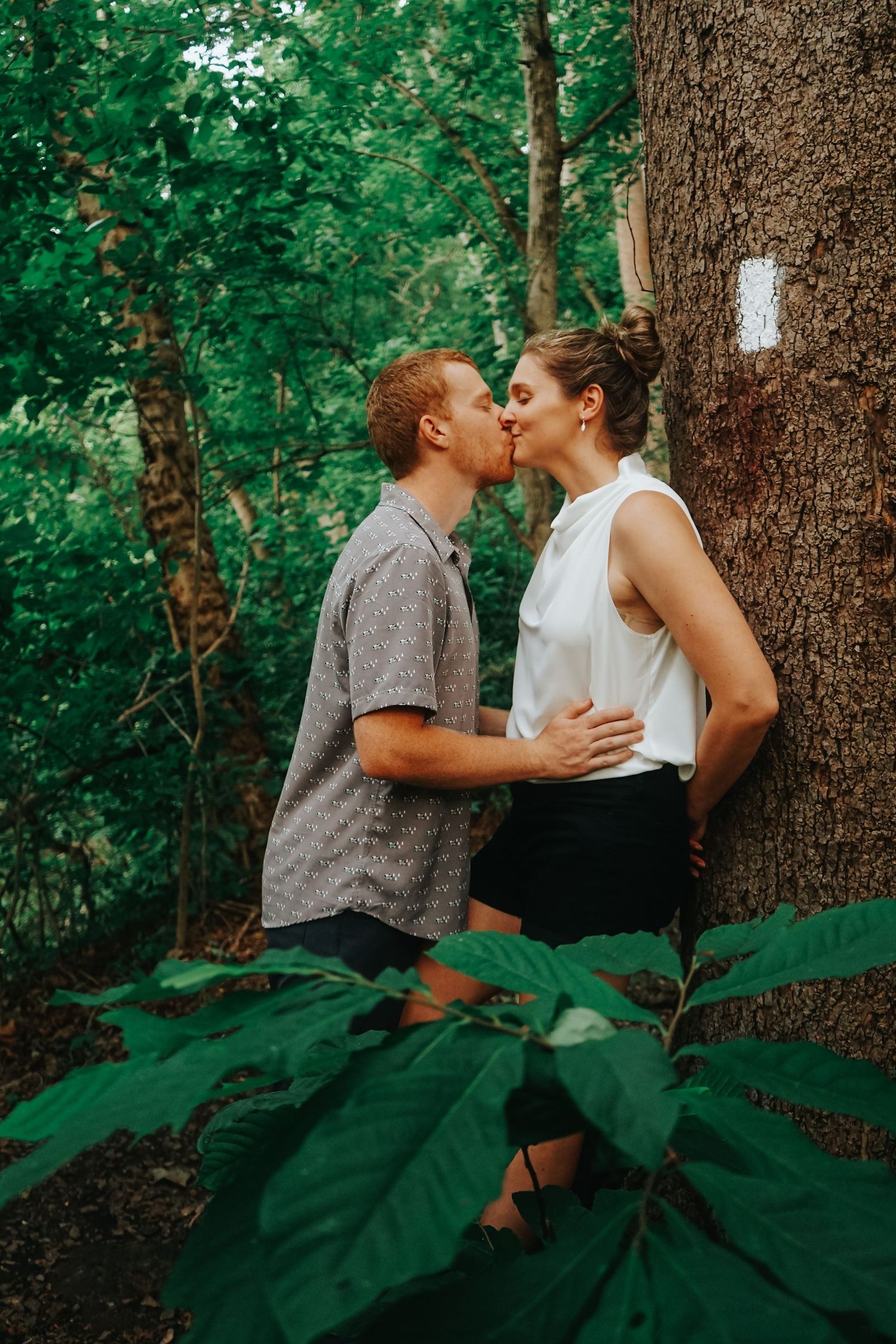 A romantic kiss shared between a couple in a lush green forest setting beside a large tree trunk. Appalachian Trail Engagement