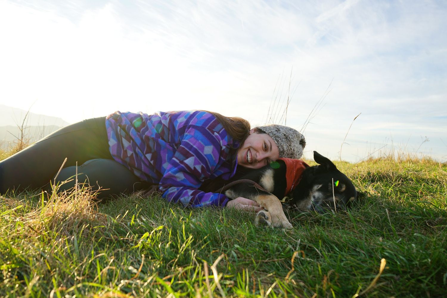 Someone in a purple patterned jacket lies in green grass next to a black and white dog on a sunny day.