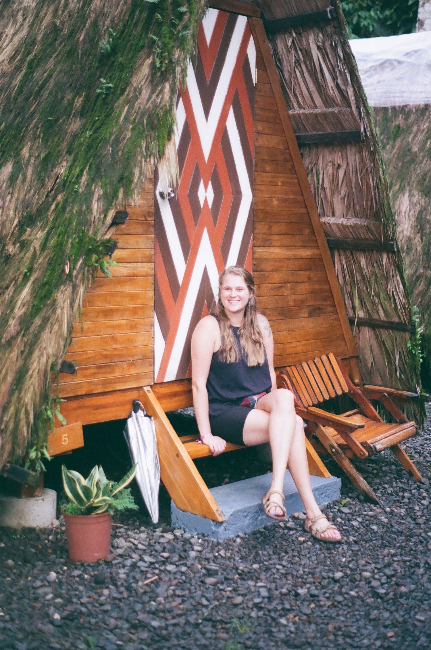 A rustic A-frame cabin with geometric wall art and wooden lounge chairs on a gravel surface.
