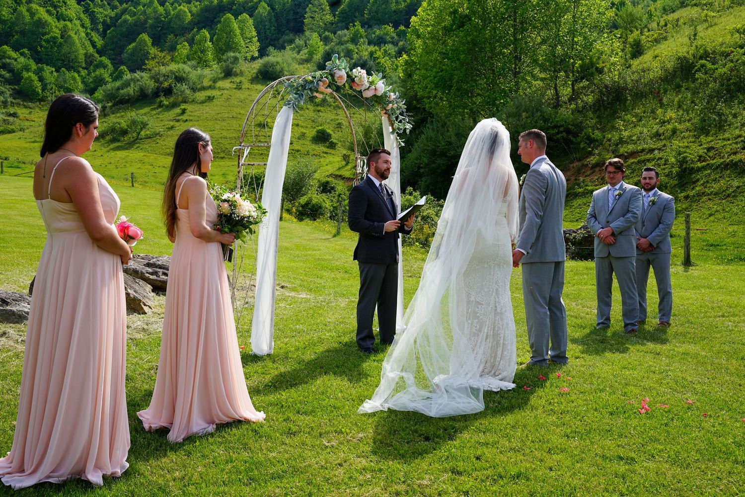 Wedding party in pink bridesmaid dresses gathers on a lush green hillside during an outdoor summer ceremony.