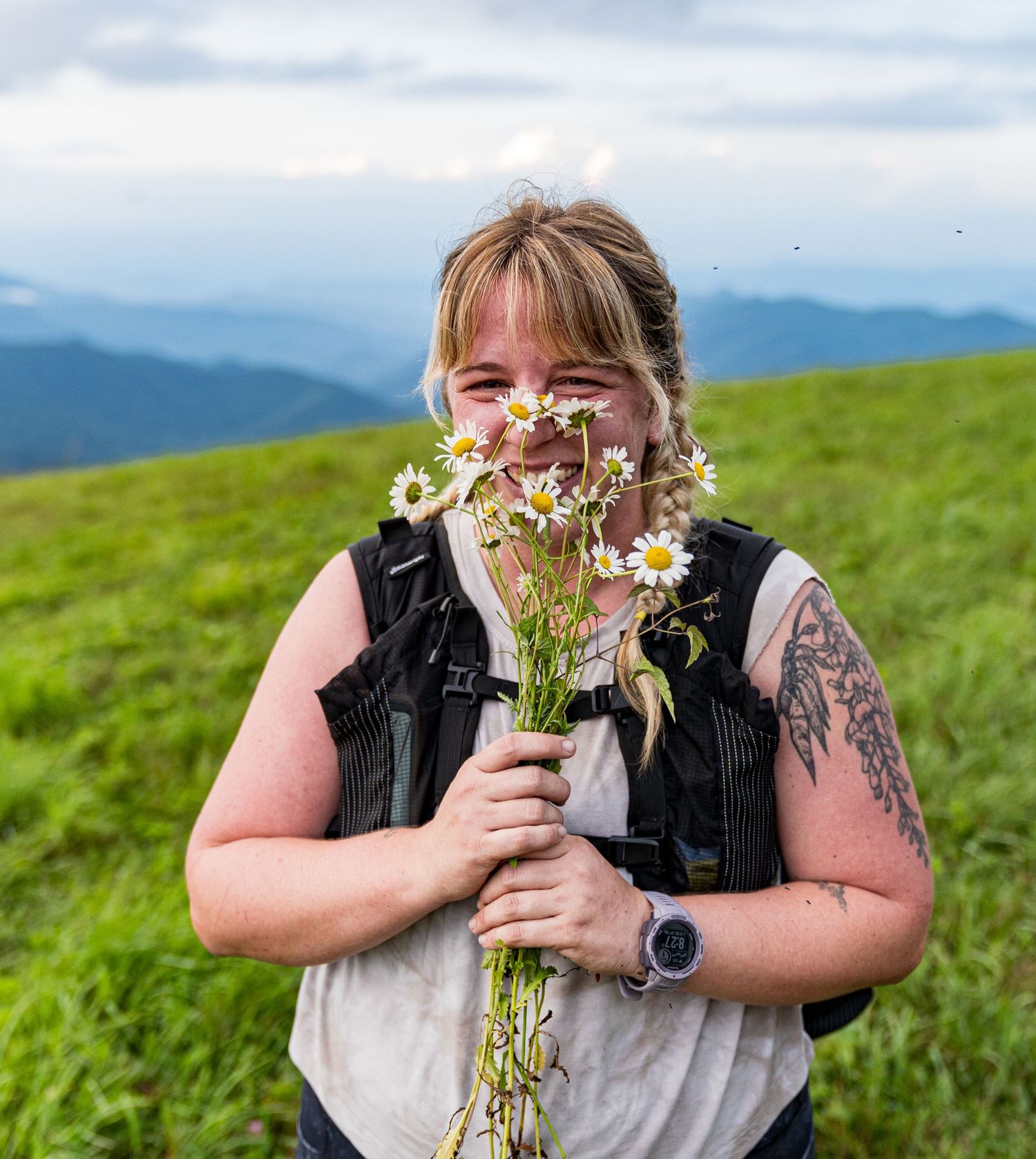 Person holding wildflowers while standing in a mountainous meadow on a sunny day.