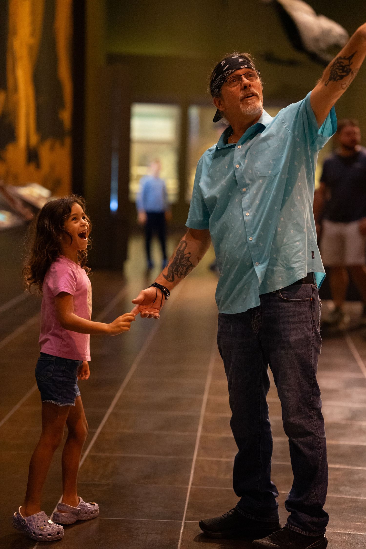 A young girl in a pink shirt and shorts reaching up to hold hands with an adult in a blue shirt in a hallway.