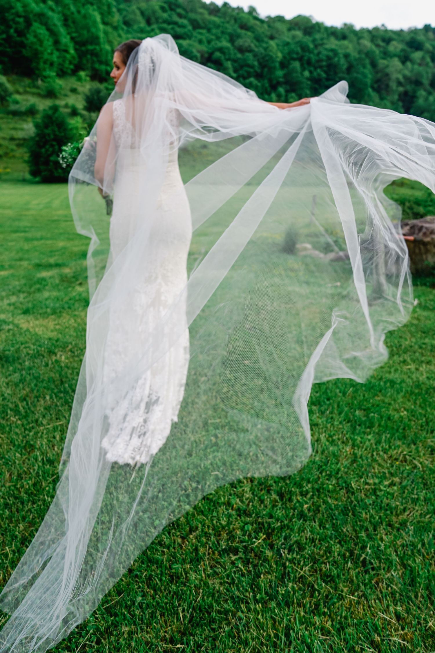 A flowing white wedding veil billows gracefully in the wind against a lush green outdoor backdrop.