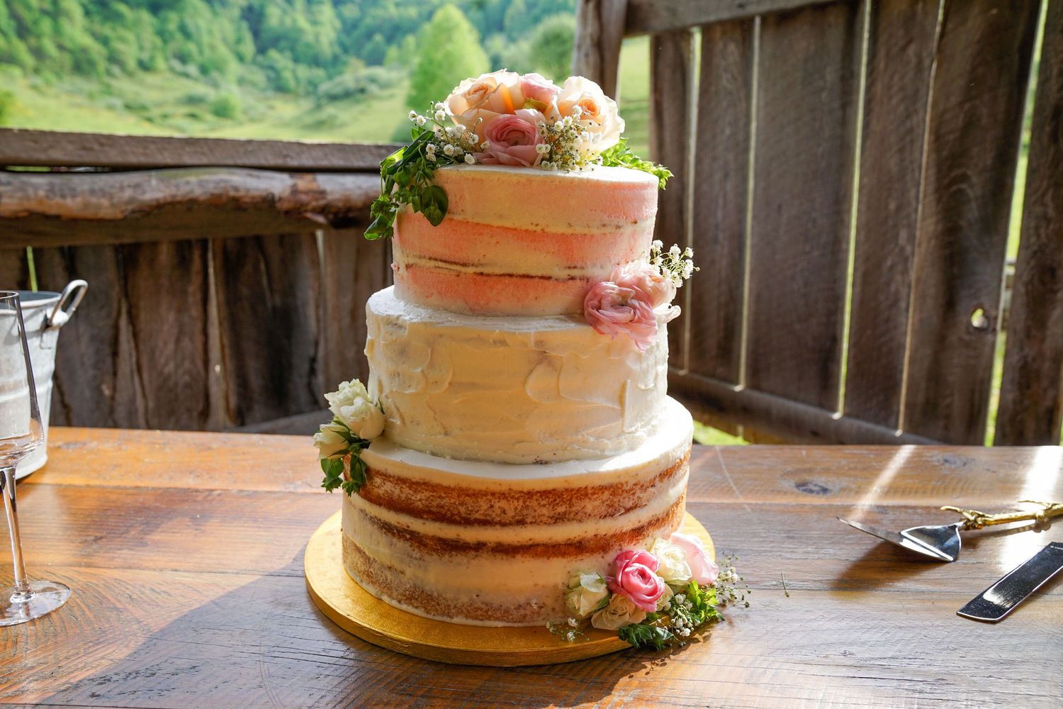 A rustic naked wedding cake decorated with fresh flowers and greenery sits on a wooden table.