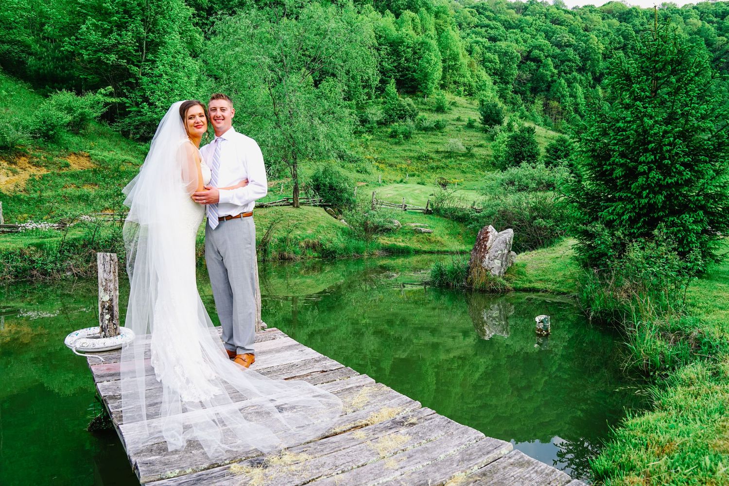 A couple in wedding attire poses together in a lush green garden setting during their summer celebration.