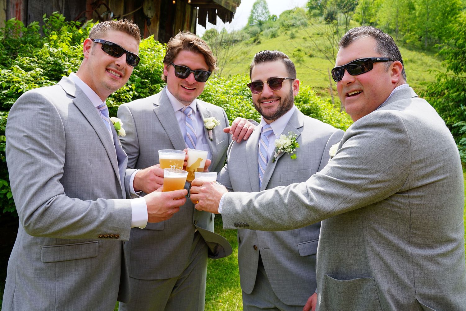 A sequence of groomsmen in gray suits toasting and clinking beer bottles together during an outdoor wedding celebration.