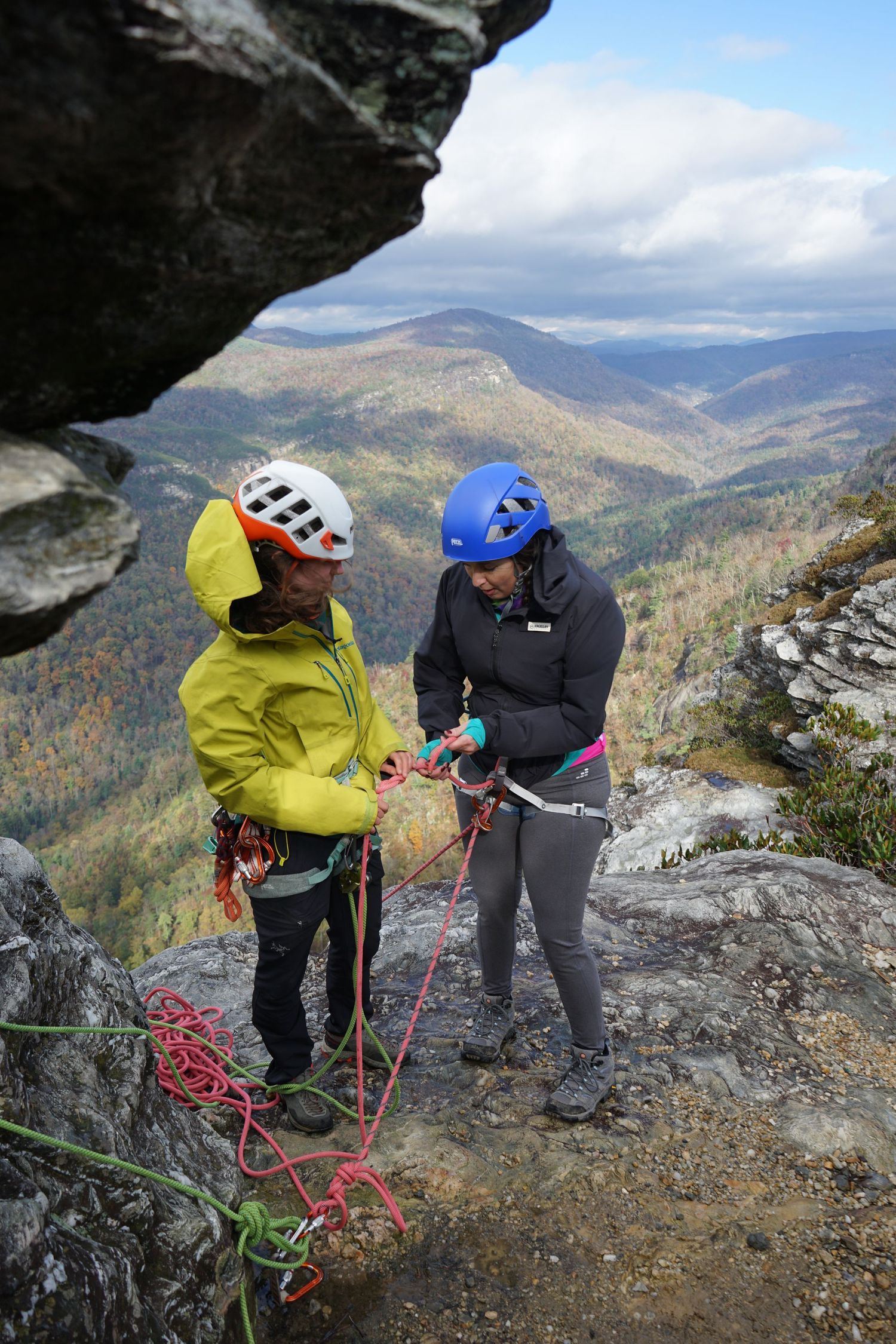 Two climbers set up ropes and gear on a rocky mountain ledge with autumn valley views below.