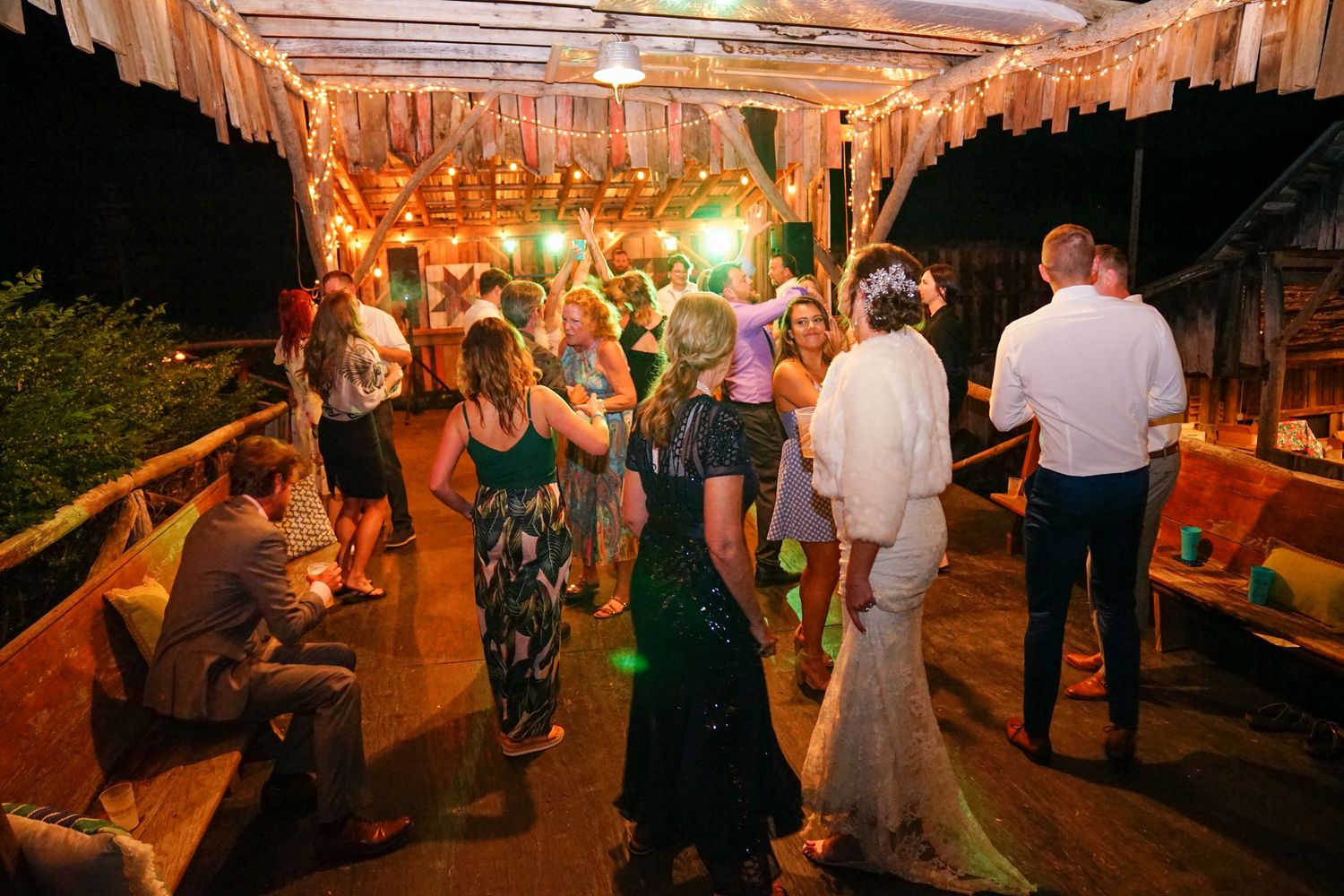 People dancing and socializing at an outdoor party venue decorated with string lights and rustic wooden beams.