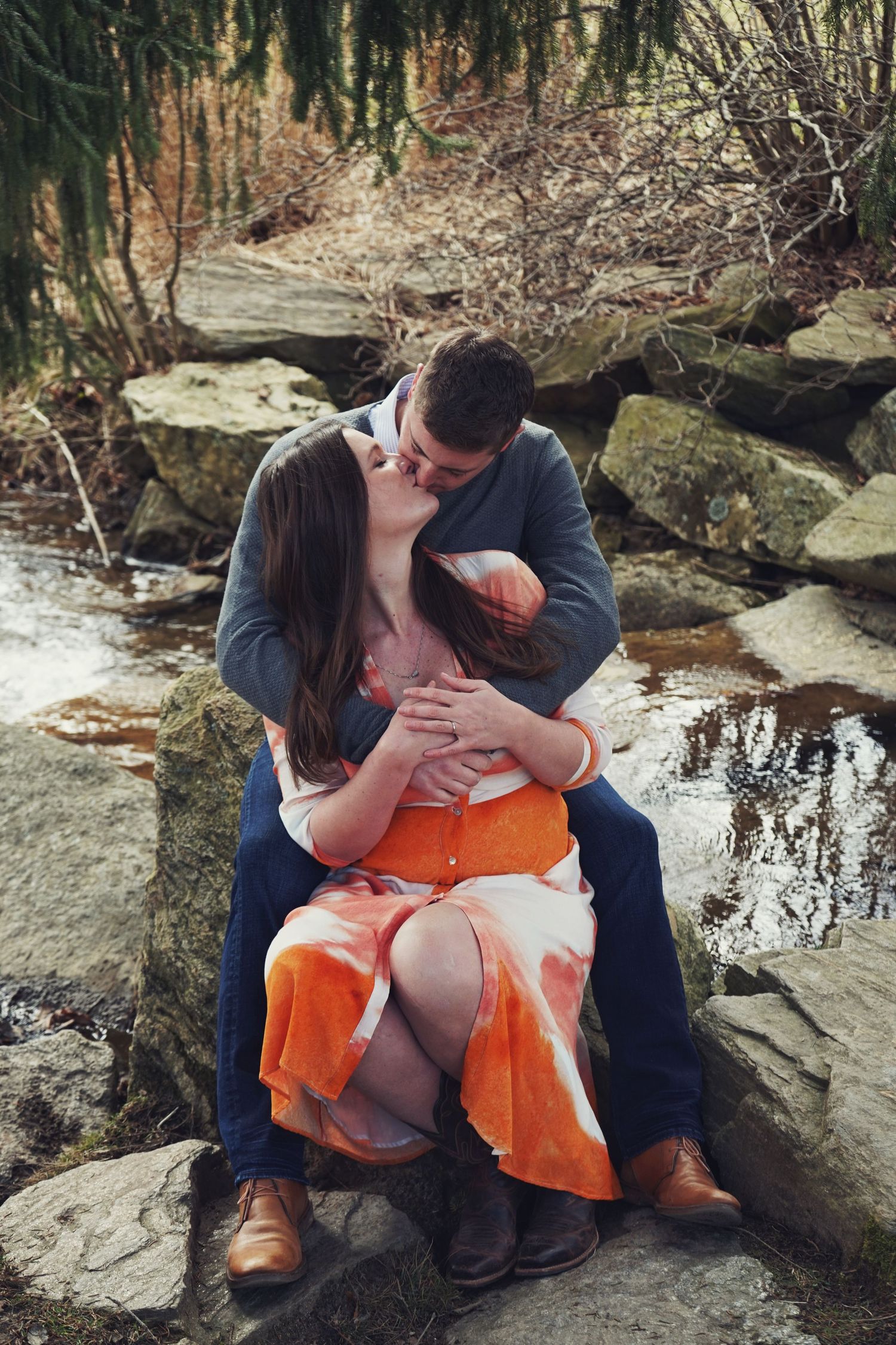 A couple shares an intimate moment sitting on rocks by a stream, with the person wearing an orange dress.