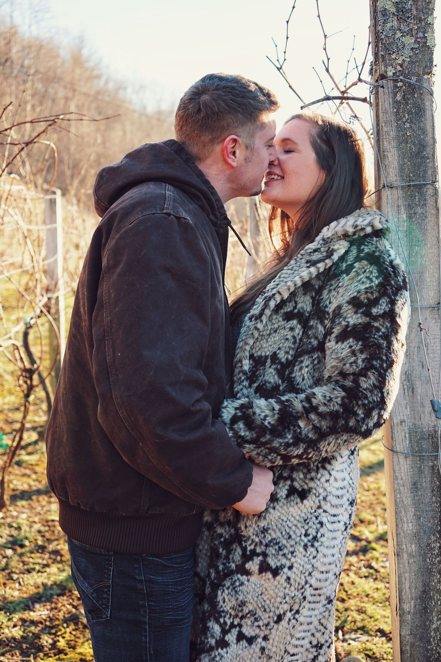A couple embraces in a winter forest scene with bare trees in the background.