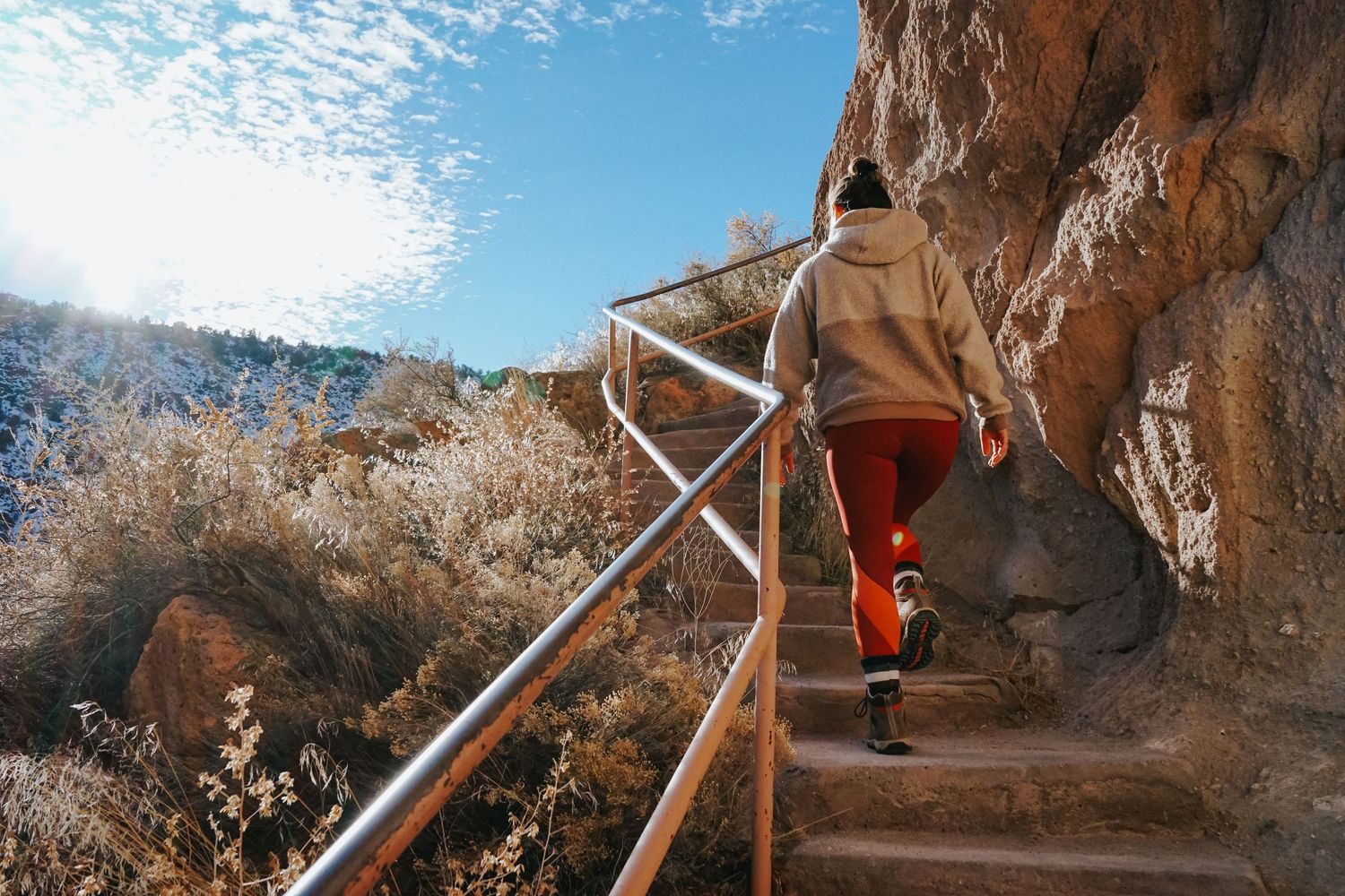A hiker in bright red pants climbs up steep stone steps with metal handrails along a cliffside mountain trail.