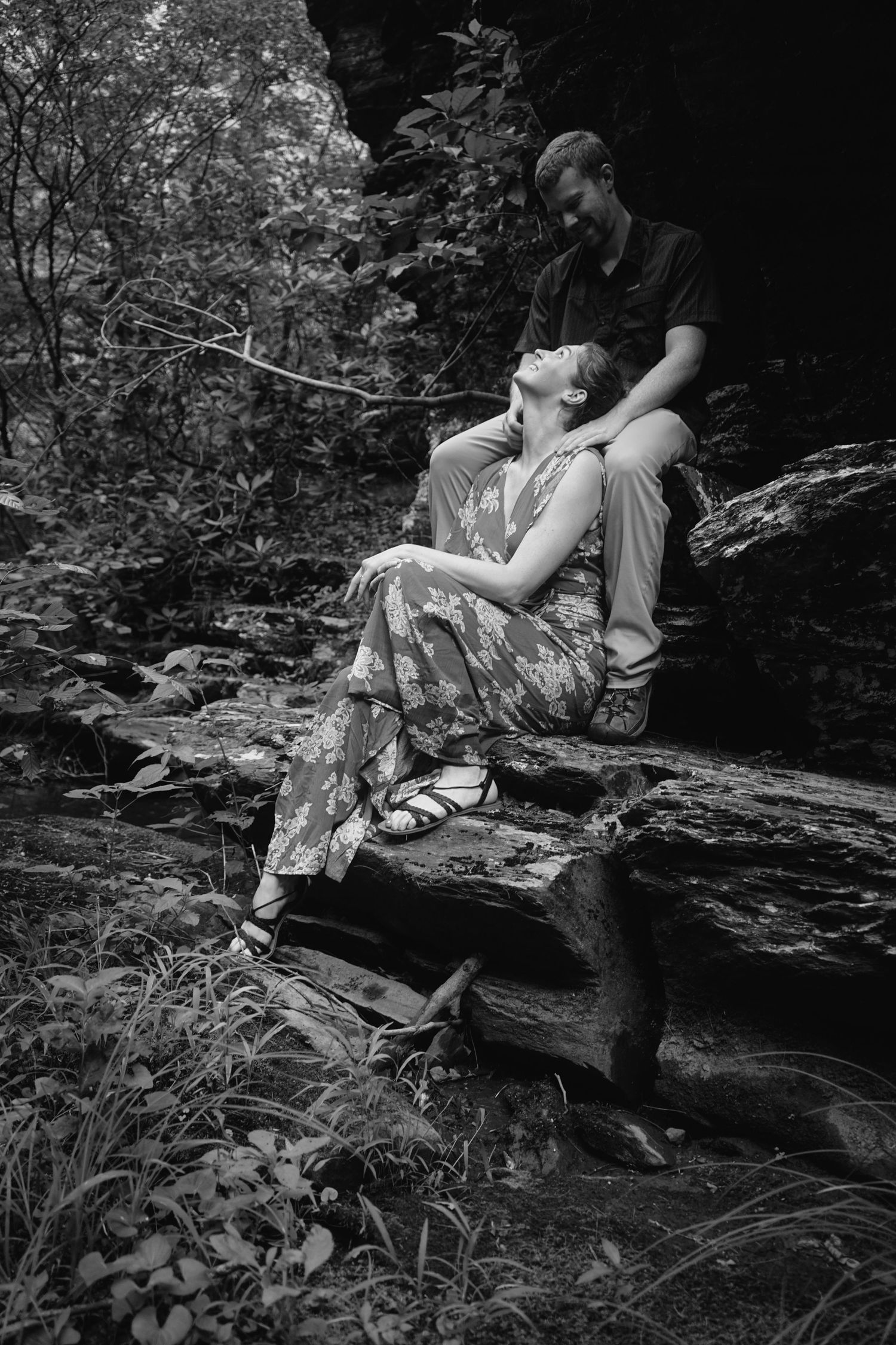 Romantic black and white photograph of a couple resting together on rocky terrain surrounded by foliage in nature.